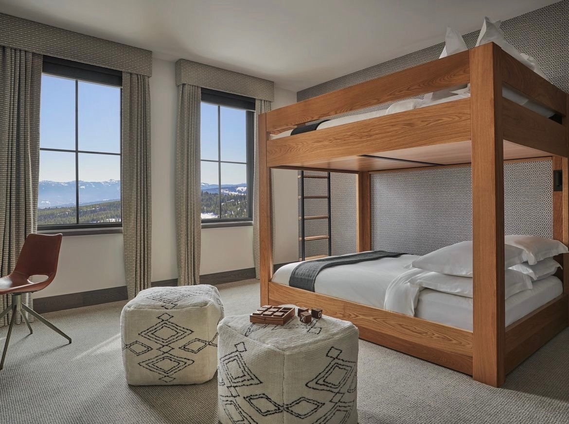 A bedroom with a wooden bunk bed, two large windows showing mountainous landscape, a brown chair, two patterned ottomans, and a small table with game pieces.