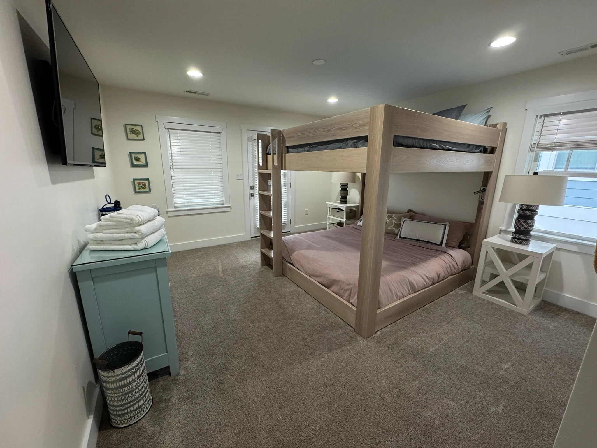 A bedroom with a bunk bed, restuffed with a ground-level bed with pink bedding and pillows, a nightstand, a lamp, window blinds, and a wall-mounted TV.