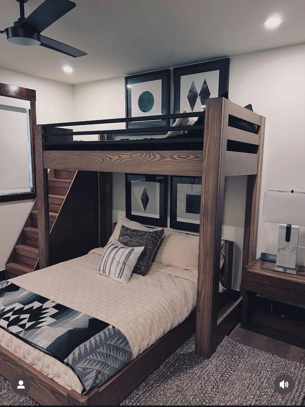 Bedroom with a wooden loft bed, a staircase, a queen-sized bed with pillows and a quilt, framed abstract art on the wall, a window, a nightstand with a lamp and a decorative item, and a ceiling fan.