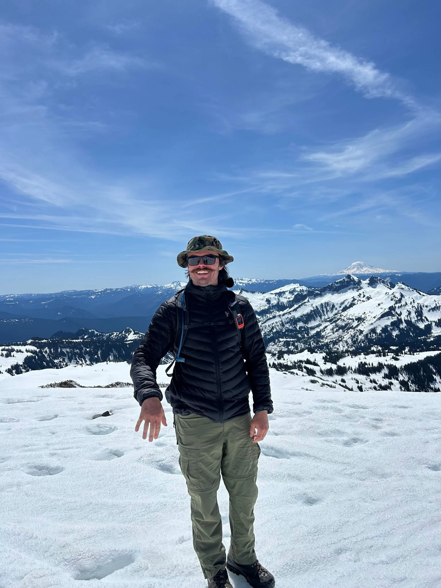 A man standing on snow-covered mountain terrain with snow-capped peaks and a blue sky with wispy clouds in the background, wearing outdoor gear including a camouflage hat, sunglasses, black jacket, and khaki pants.