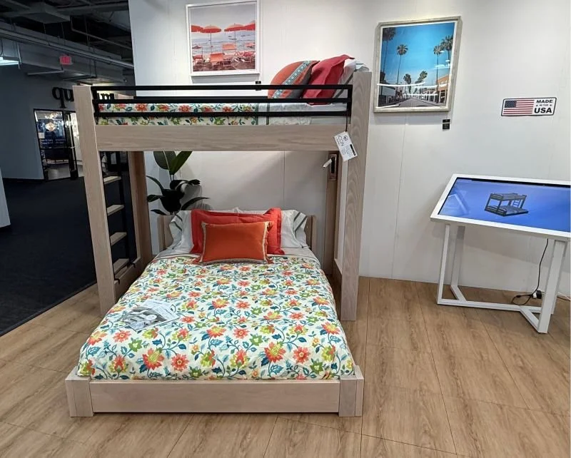 Bunk bed with floral bedding, orange and red pillows, located in a furniture store. Framed pictures of beaches and palm trees on the wall, a plant nearby, and a touchscreen display table on the right. Small American flag sign on the wall.