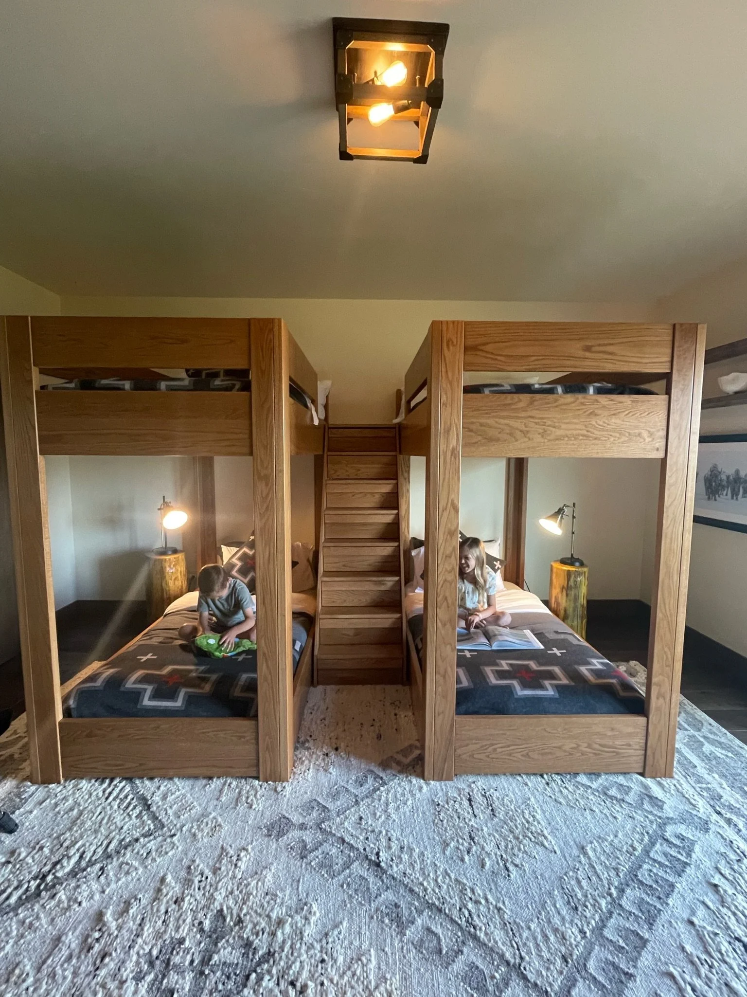 Two children playing on separate beds below a wooden bunk bed structure in a bedroom with a patterned rug, side lamps, and a ceiling light.