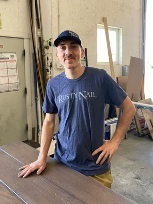 Man wearing a blue Rusty Nail t-shirt standing in a workshop with wood and tools in the background.