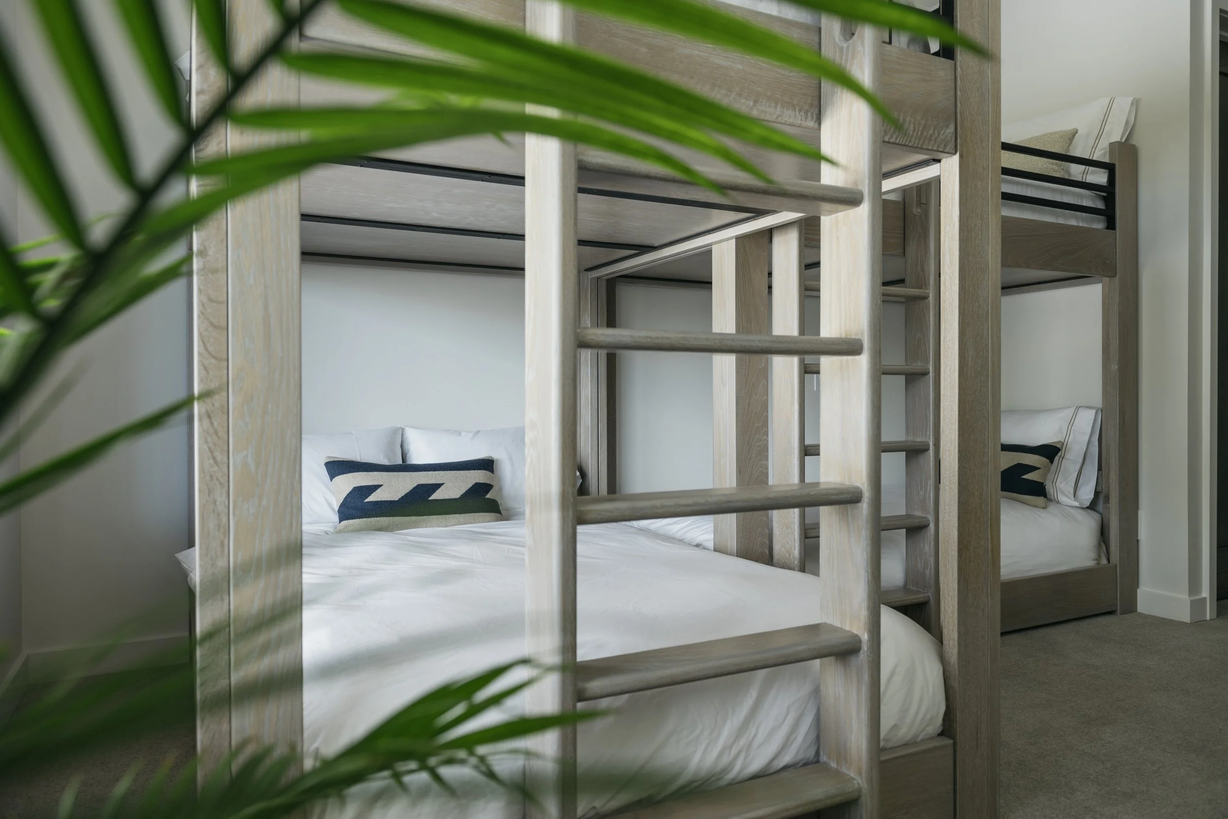 Bunk beds with wooden frames in a bedroom, viewed through green leaves.