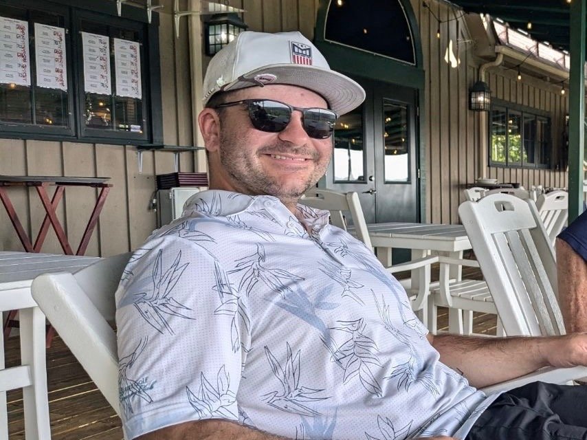 A man wearing sunglasses, a white cap, and a light-colored Hawaiian shirt with a leaf pattern, smiling and sitting outdoors at a restaurant or cafe.