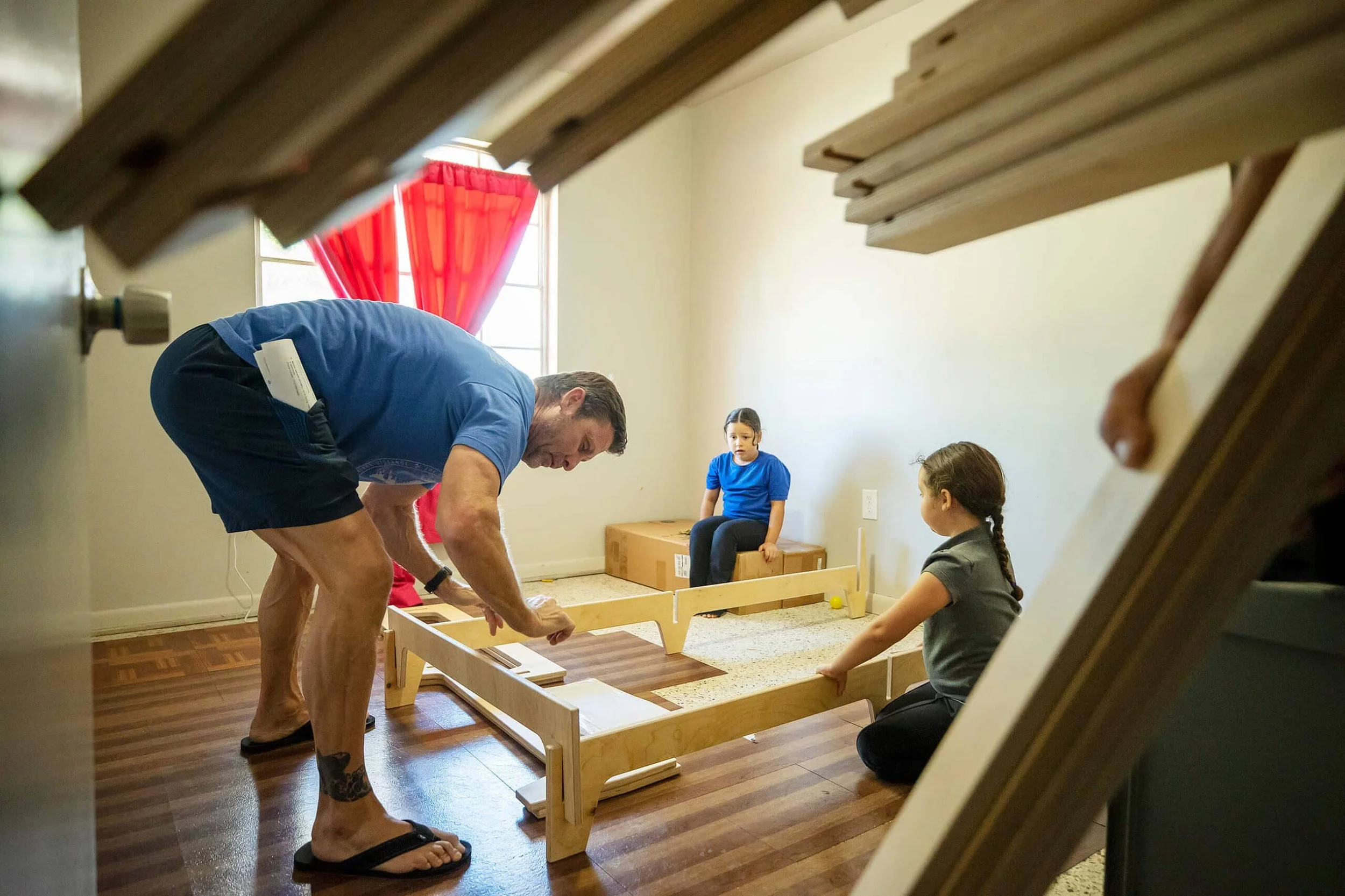 A man and two young girls working on building a miniature wooden roller coaster in a room with hardwood floors and a window with red curtains.
