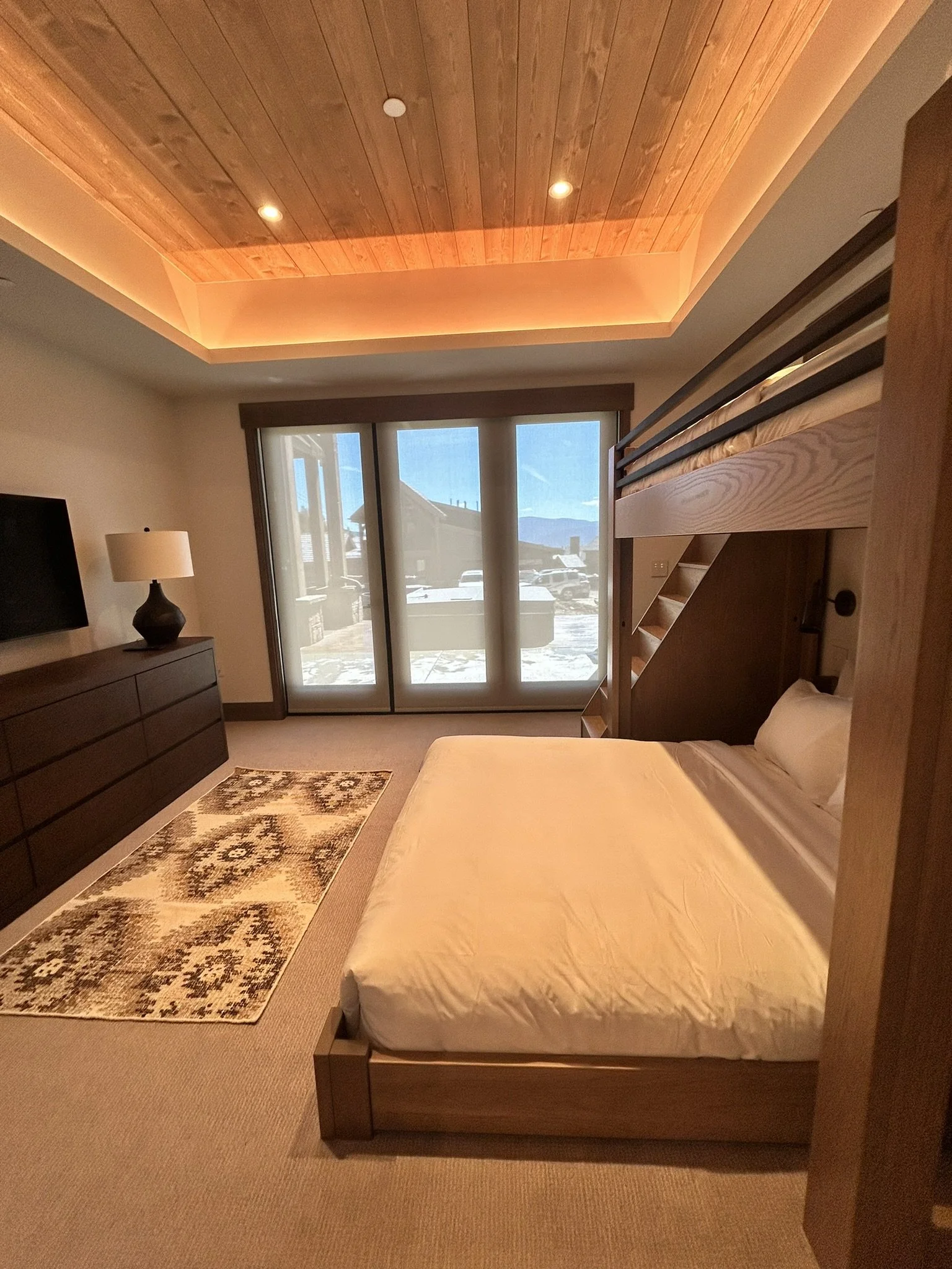 Bedroom with a loft bed, a sliding glass door, a TV on a dresser, a lamp, and a patterned rug.