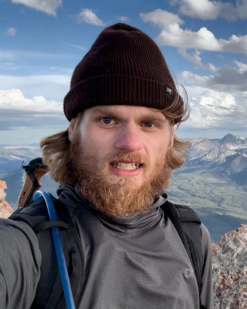 A man with a beard and long hair wearing a beanie and hiking gear taking a selfie on a mountain with a scenic background of mountains and a partly cloudy sky.