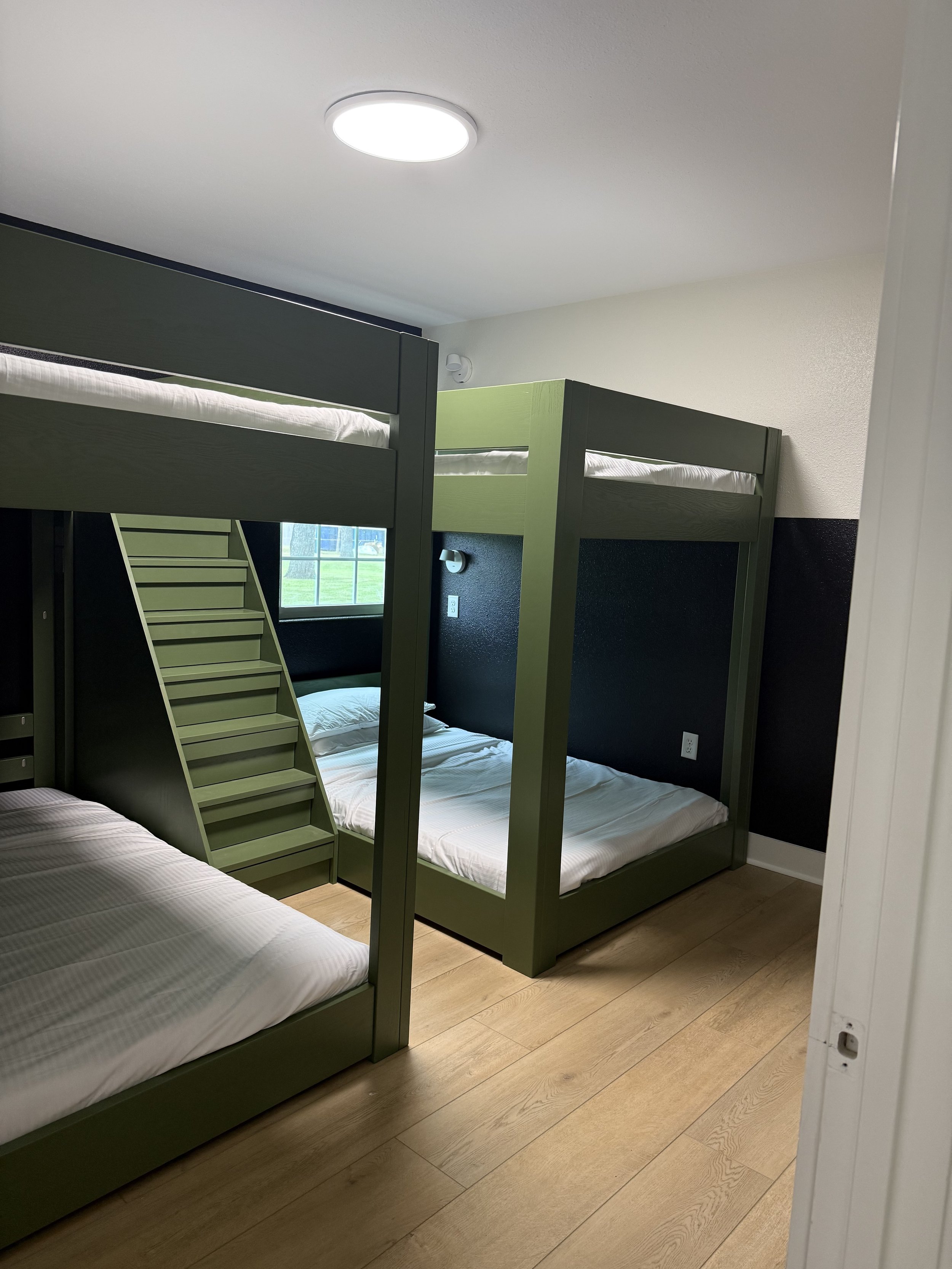 Room with three green bunk beds and a window showing grass outside.