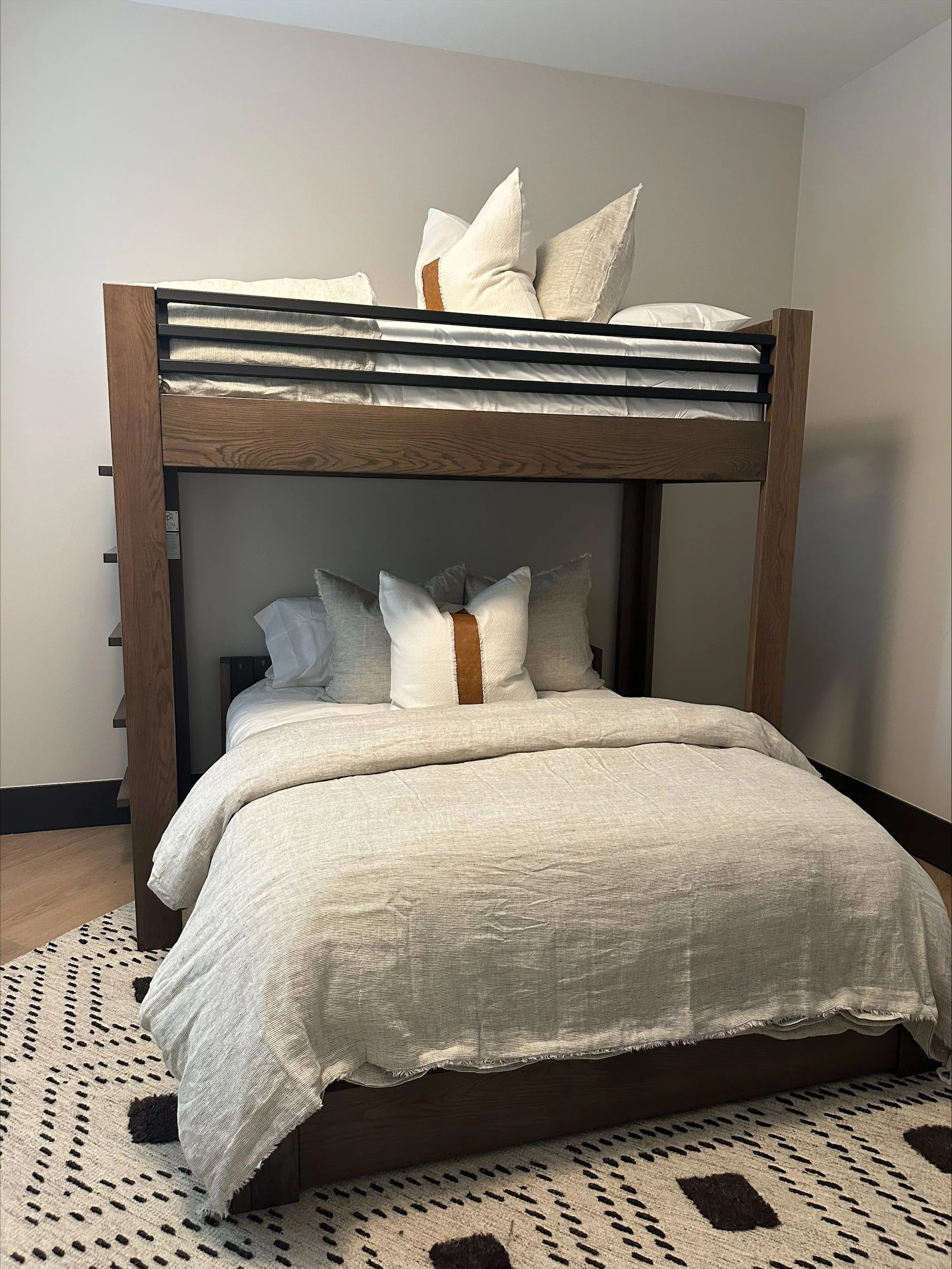 A bedroom setup with a bunk bed featuring a lower double bed and an upper twin bed, both with beige bedding and pillows, positioned against plain white walls on a patterned rug.
