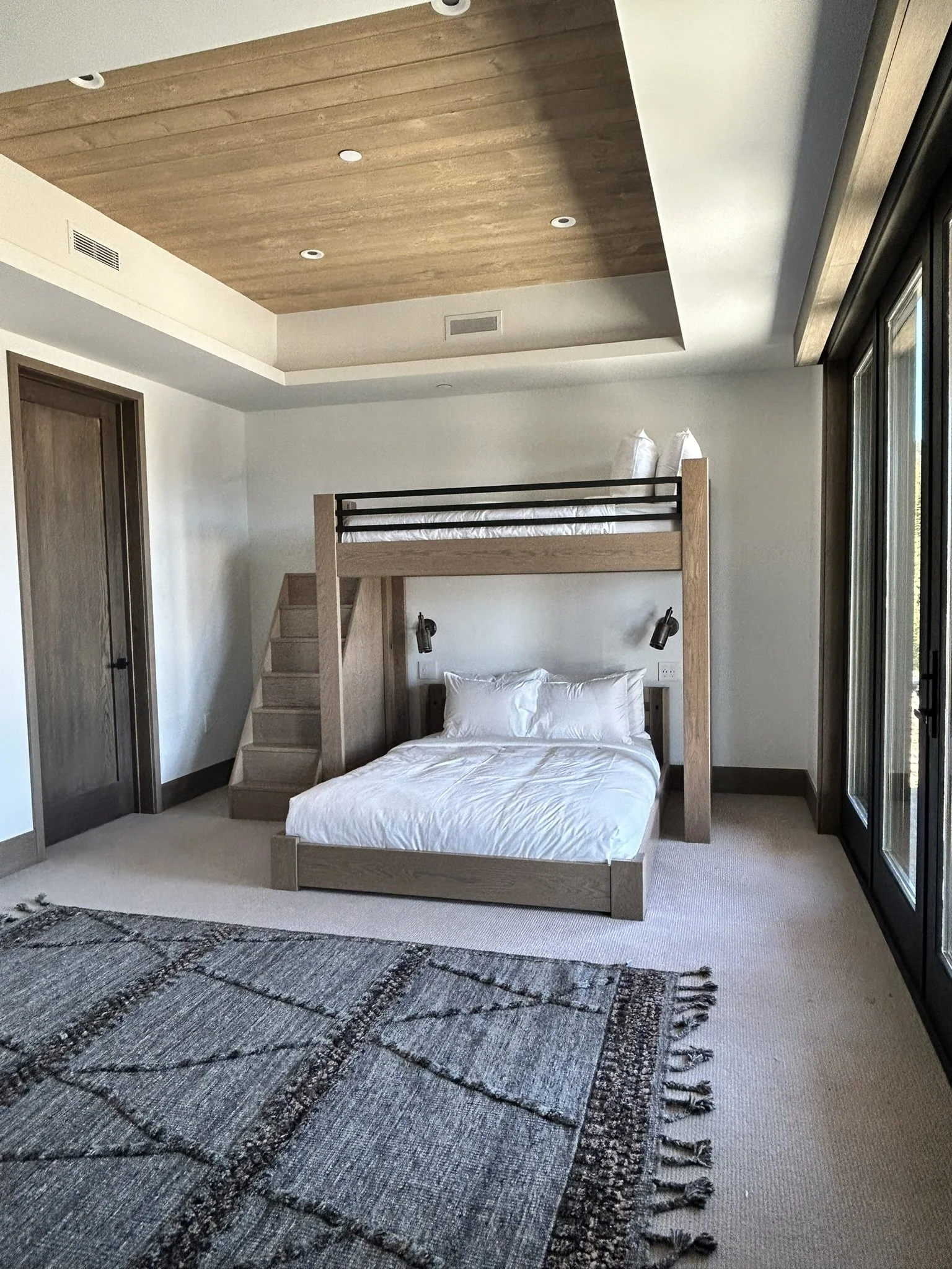 A bedroom with a bunk bed where the bottom is a double bed and the top bed is a twin, with sliding glass doors on the right and a patterned rug on the beige carpeted floor.