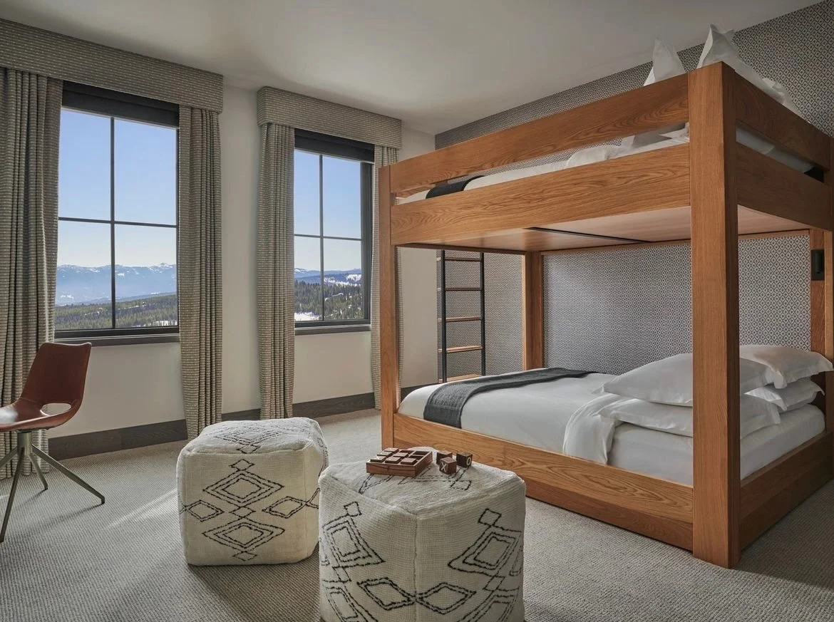 A bedroom with two large windows showing a mountain view, a wooden bunk bed with a black ladder, a white chair, and two patterned ottomans.
