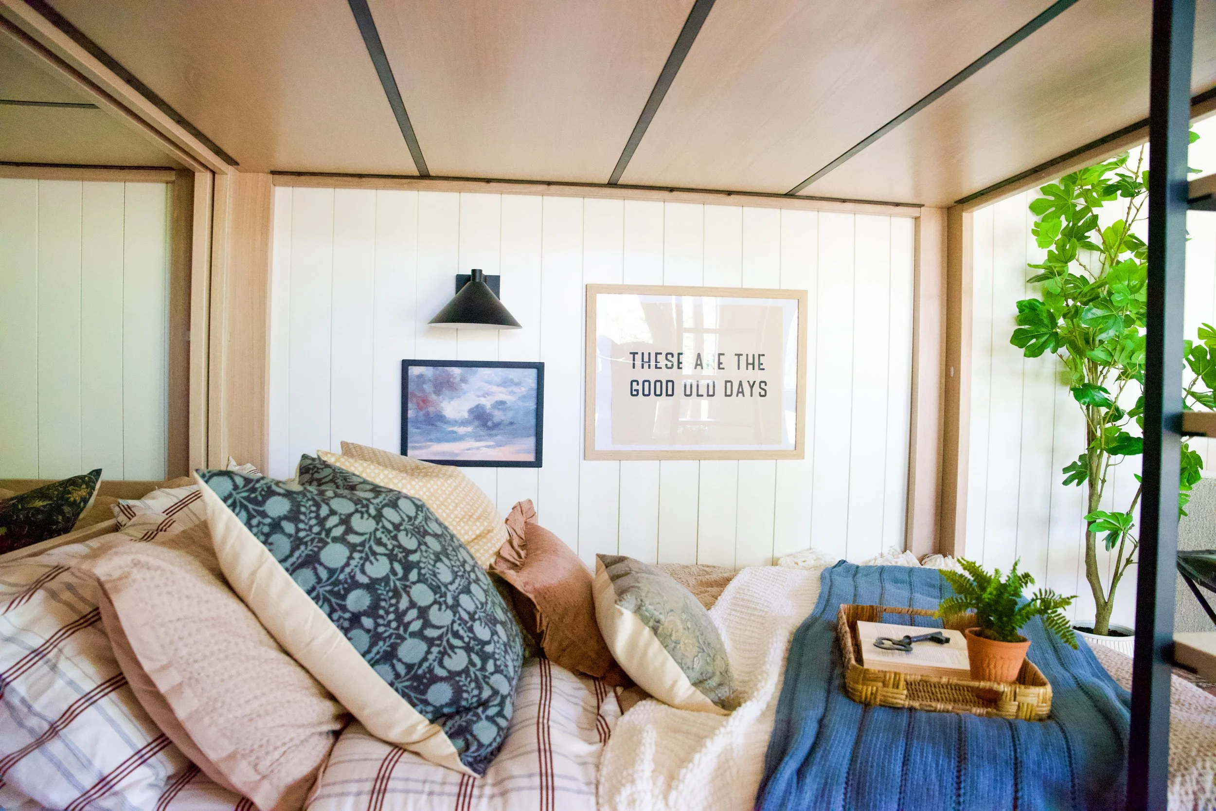 A cozy bedroom with a bed covered in various pillows and blankets, a small wicker tray with a plant on a blue throw, a framed wall art reading 'These Are The Good Old Days', a picture of the sky, a black wall lamp, and a large green plant near the wi