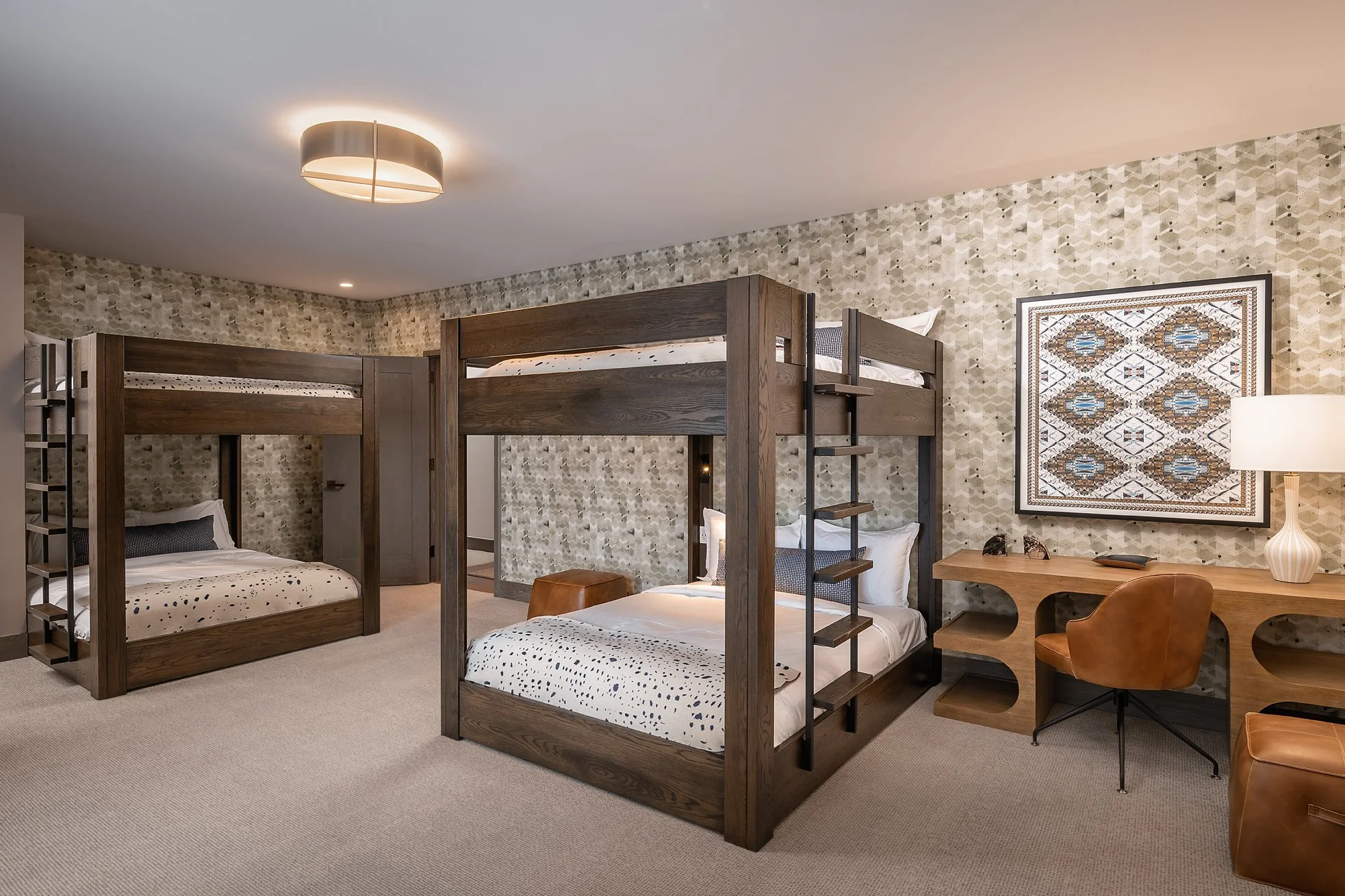 A hotel room with two bunk beds, a desk with a chair, a table lamp, and a framed decorative artwork on the wall.