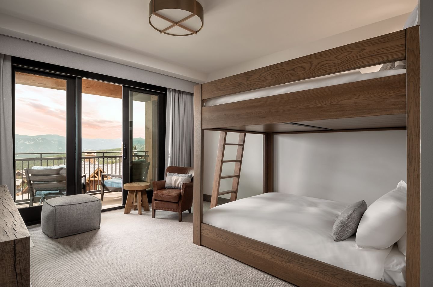 Hotel room with bunk bed, sitting area with armchair, ottoman, small table, and balcony with mountain view.
