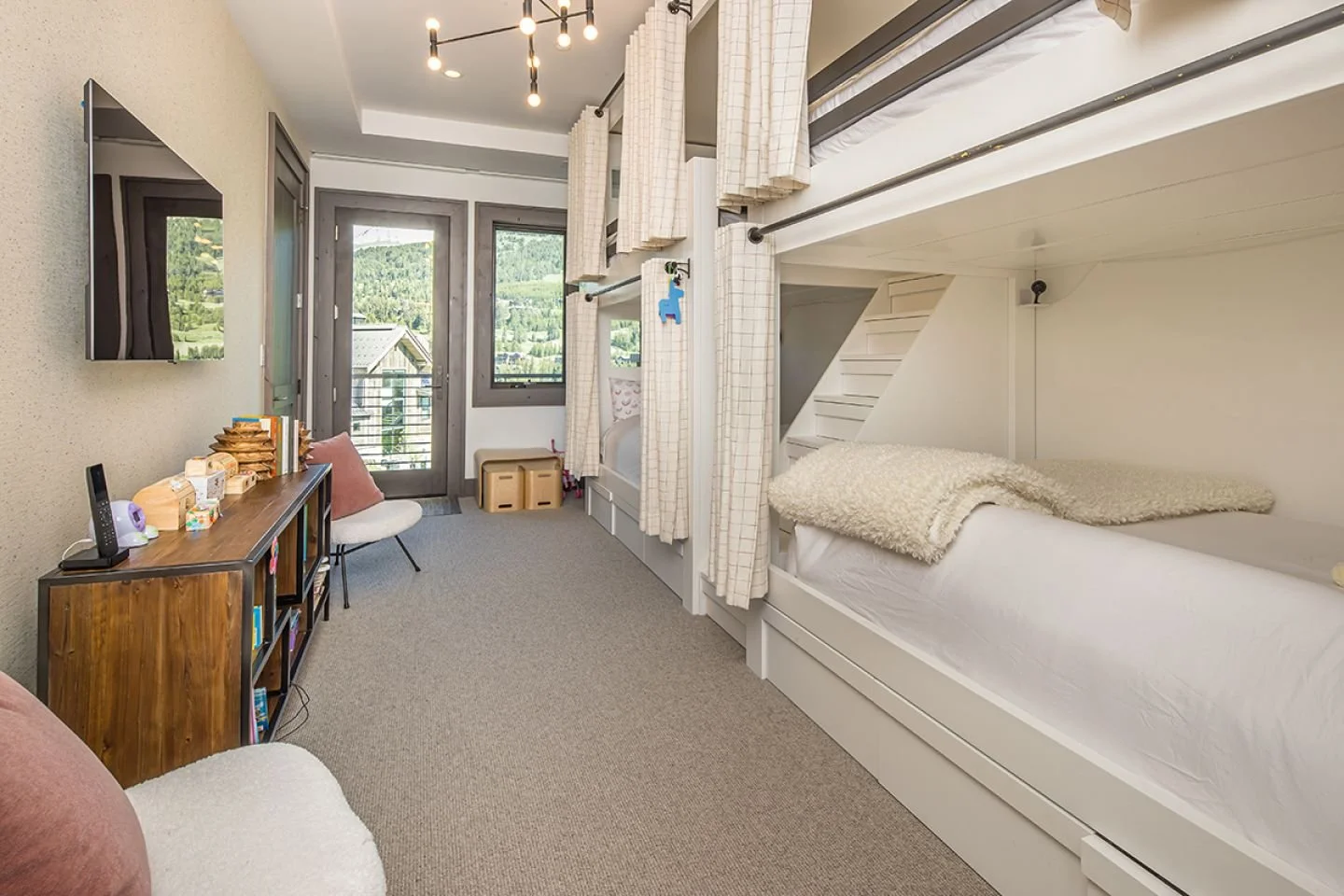 A cozy bedroom with a bunk bed, small sofa, TV on wall, wooden console table, and balcony door with mountain view.