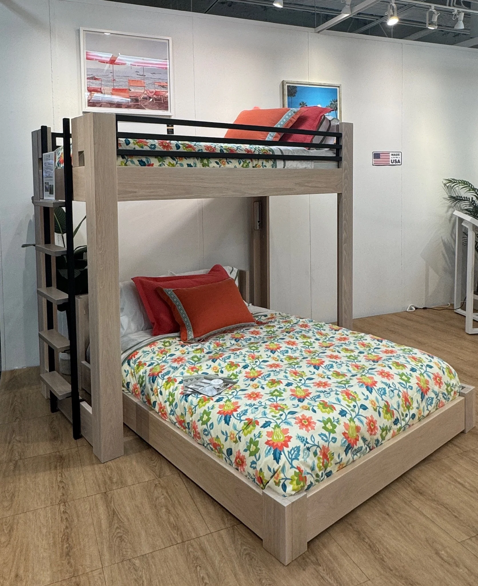 A bunk bed with a floral-patterned bedspread on the bottom and a colorful pillow. The top bunk has an orange pillow and a multicolored bedspread. There are framed beach-themed photos hanging on the white wall behind the beds, and a small plant and la