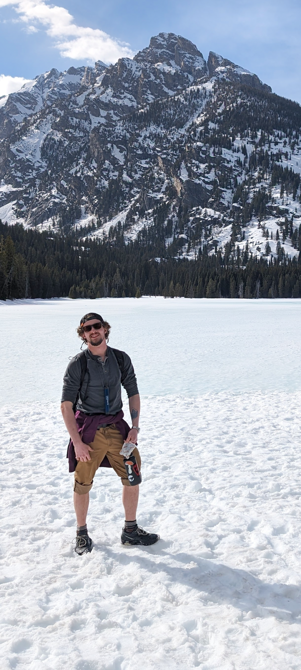A man in sunglasses and outdoor clothing kneeling on a snow-covered lake with a mountain range and forest in the background.