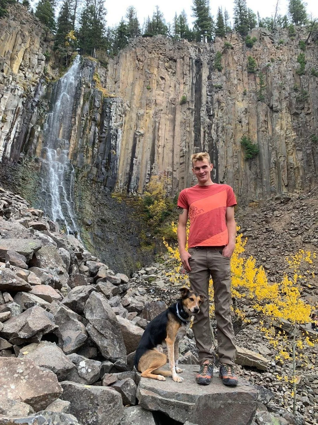 A young man in a red and orange t-shirt and khaki pants standing on rocks with a dog sitting beside him, in front of a tall cliff with a waterfall and trees in the background.