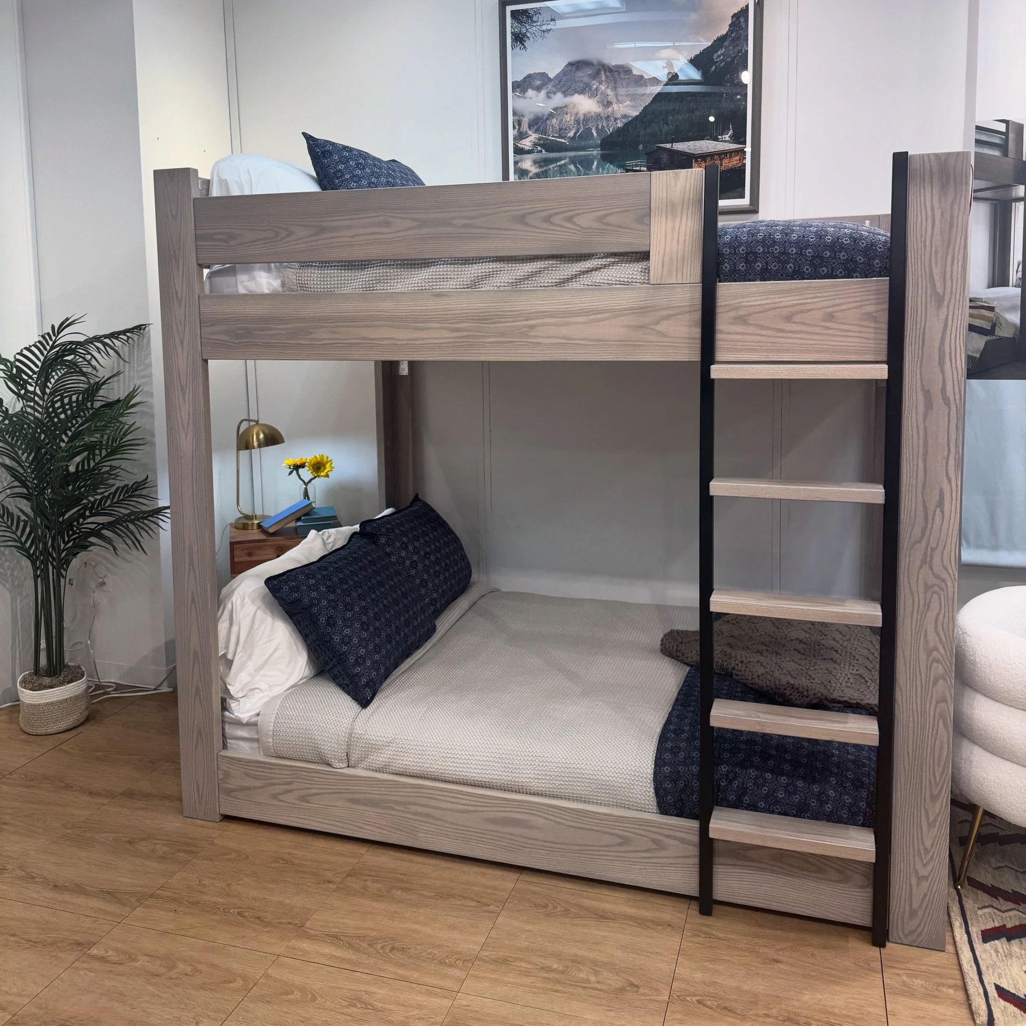 A bedroom with a wooden bunk bed, navy bed sheets, and pillows. A nightstand with a lamp, books, and a vase with yellow flowers. A framed mountain landscape picture on the wall. A potted plant near the bed.