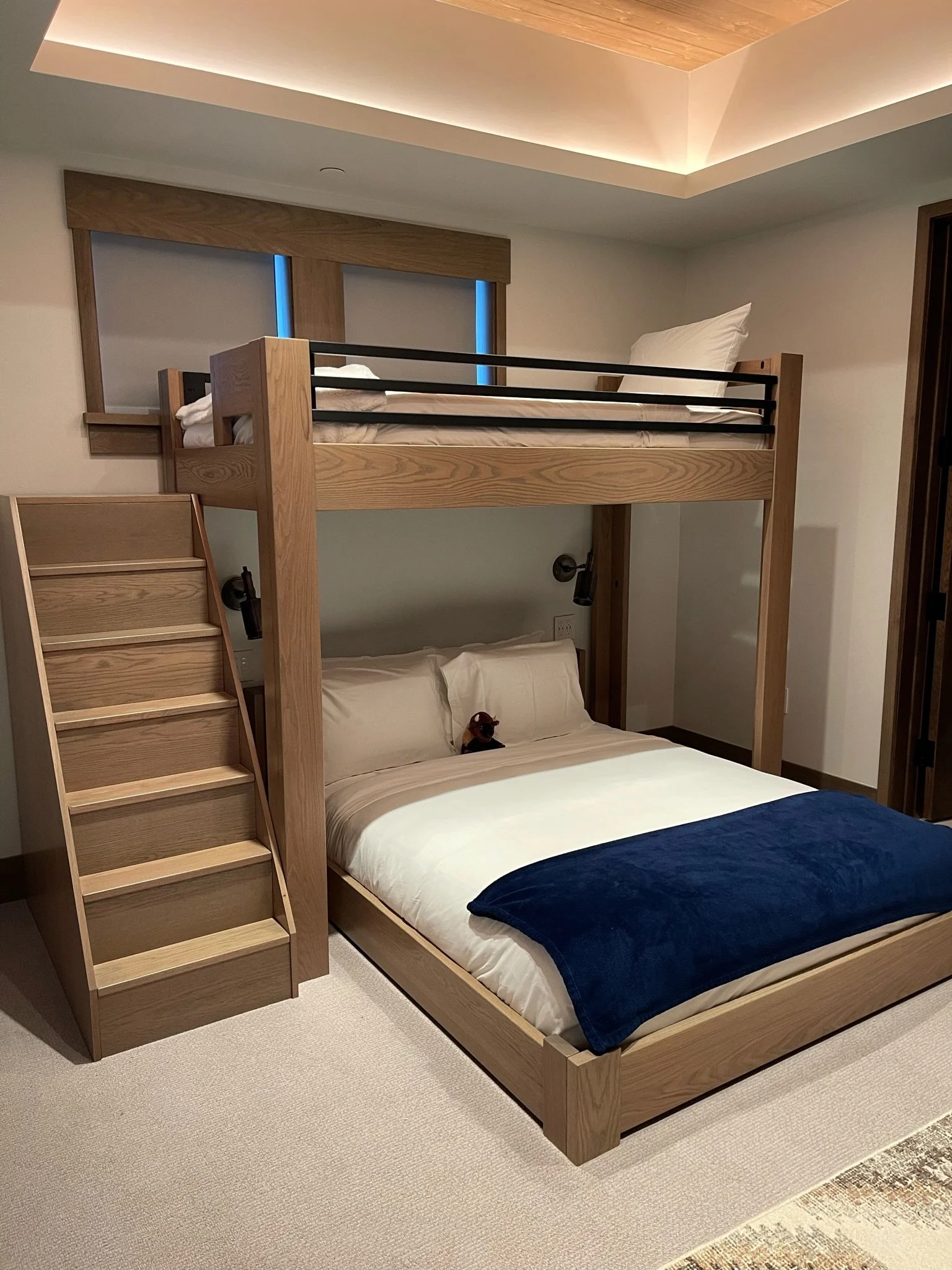 A bedroom with a wooden bunk bed that has a ladder, a lower bed with white bedding and pillows, and an upper bed with a pillow and blanket. The room has ceiling lighting, a window with wooden framing, and wall-mounted reading lights.