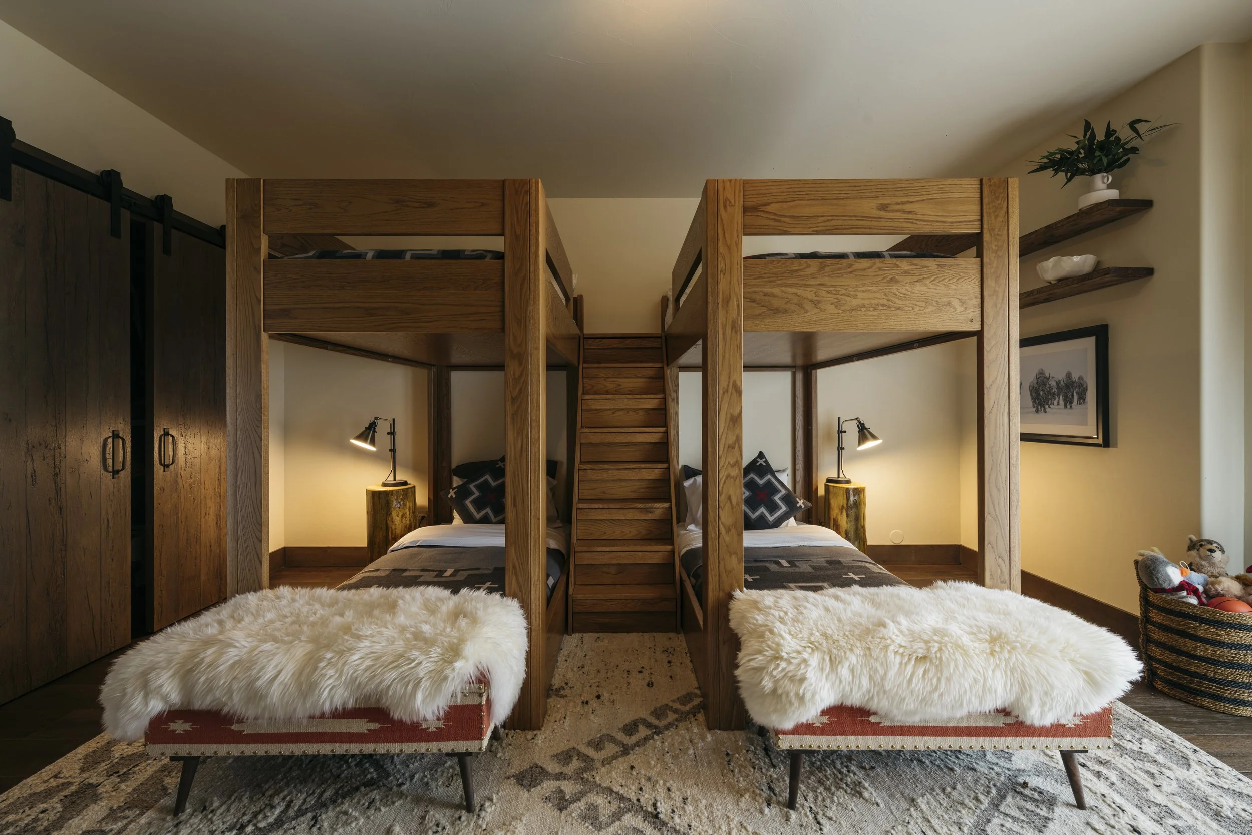 A cozy bedroom with two sets of wooden bunk beds, each with a lower bed made up with black and gray bedspreads. There are decorative pillows with geometric patterns on each bed. The room has warm lighting from two small black reading lamps attached t