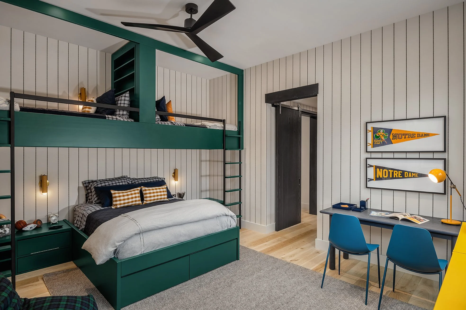 Bedroom with green bunk bed, white-paneled walls, black ceiling fan, desk with blue chairs, Notre Dame posters, and wooden floor.