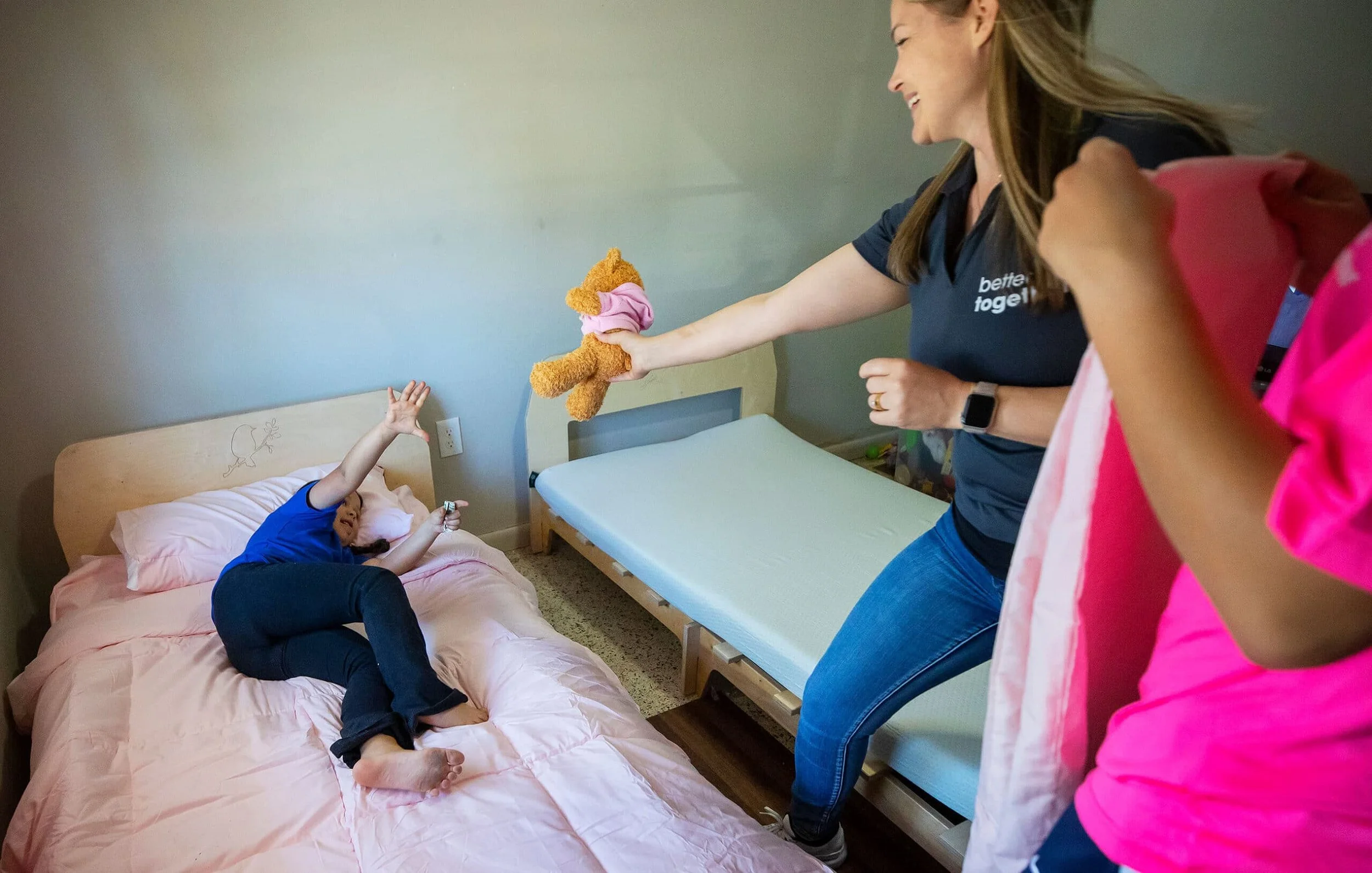 A young girl is on a bed reaching up with one hand while an adult woman, smiling, holds a stuffed dog toy towards her. The scene appears joyful and playful, in a bedroom with a second bed and neutral-colored walls.