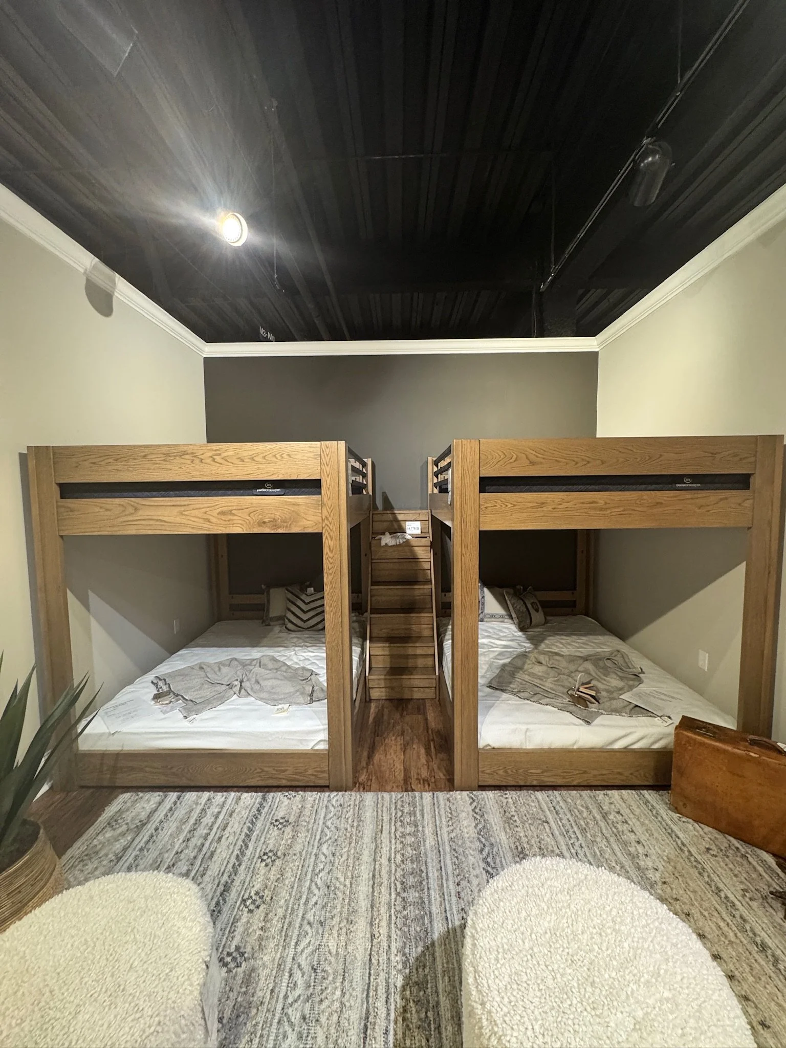 Room with two wooden bunk beds, each with a mattress, pillow, and blanket, separated by a wooden staircase, with a dark wood ceiling, light-colored walls, and a patterned rug on the floor.