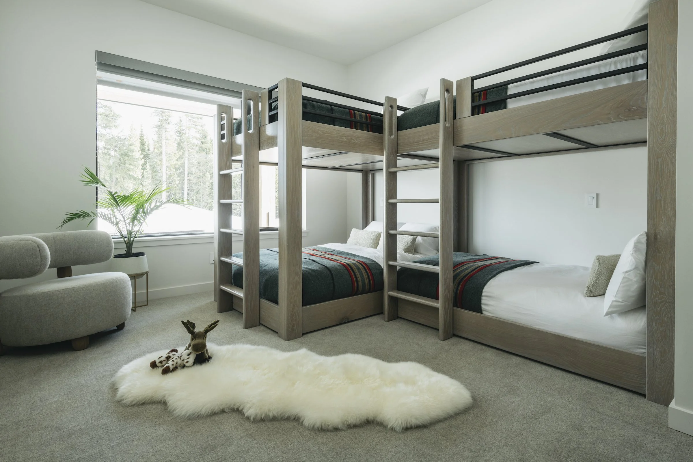 A bedroom with two sets of stacked beds, a plush white rug with a stuffed deer toy, a beige armchair, a small side table, and a large window showing trees outside.