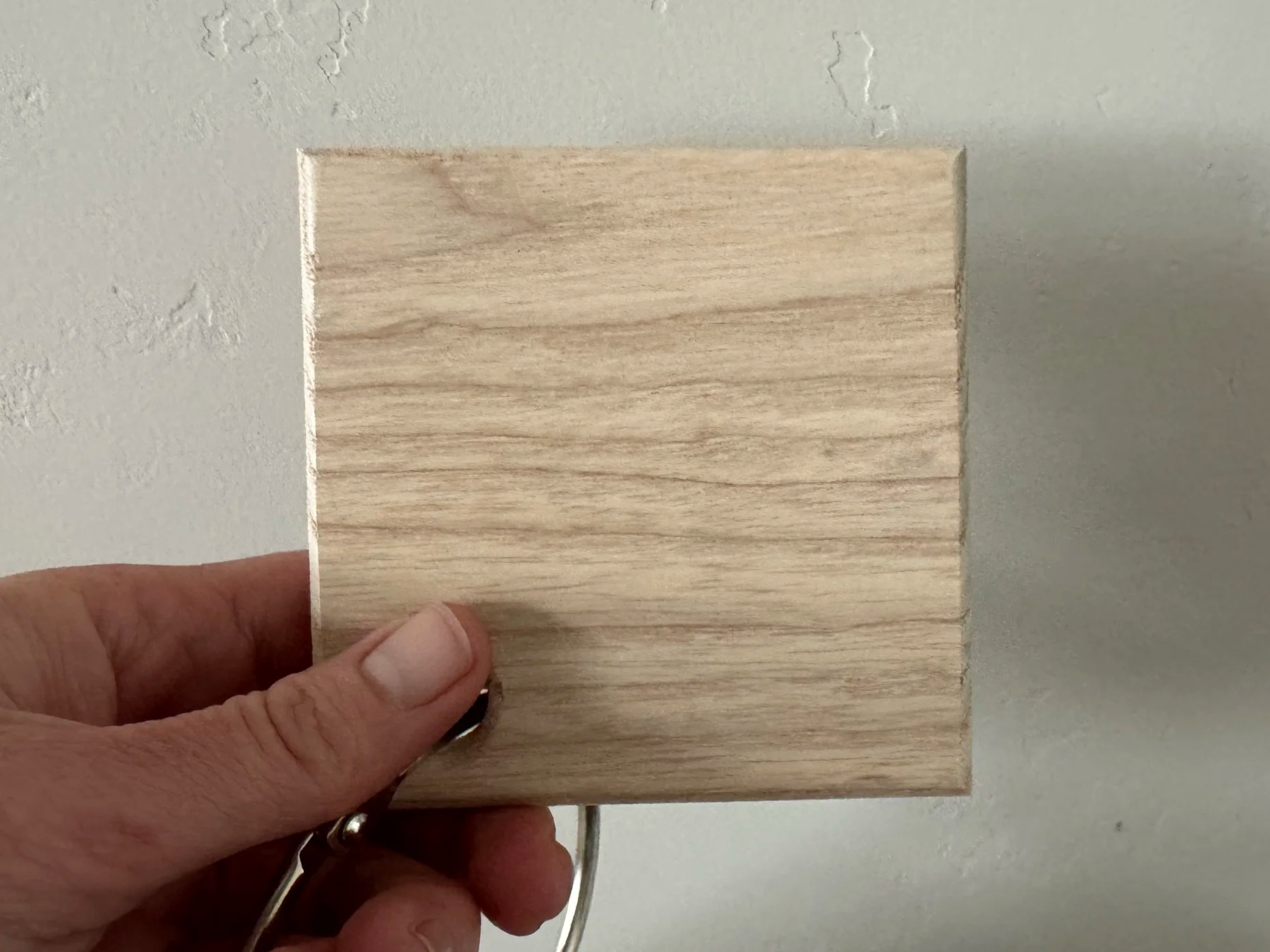 A hand holding a small, light-colored wooden block against a white wall with some texture.