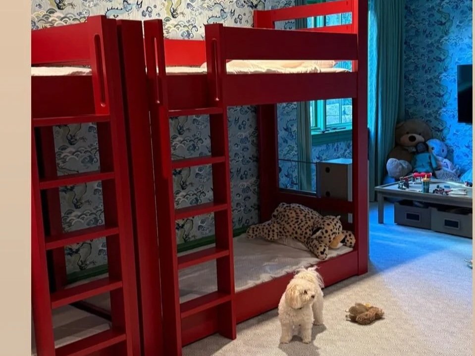 A children's bedroom with a red wooden bunk bed, a small white dog, and toys including a plush leopard lying on the lower bunk bed and a teeter-totter in the background.