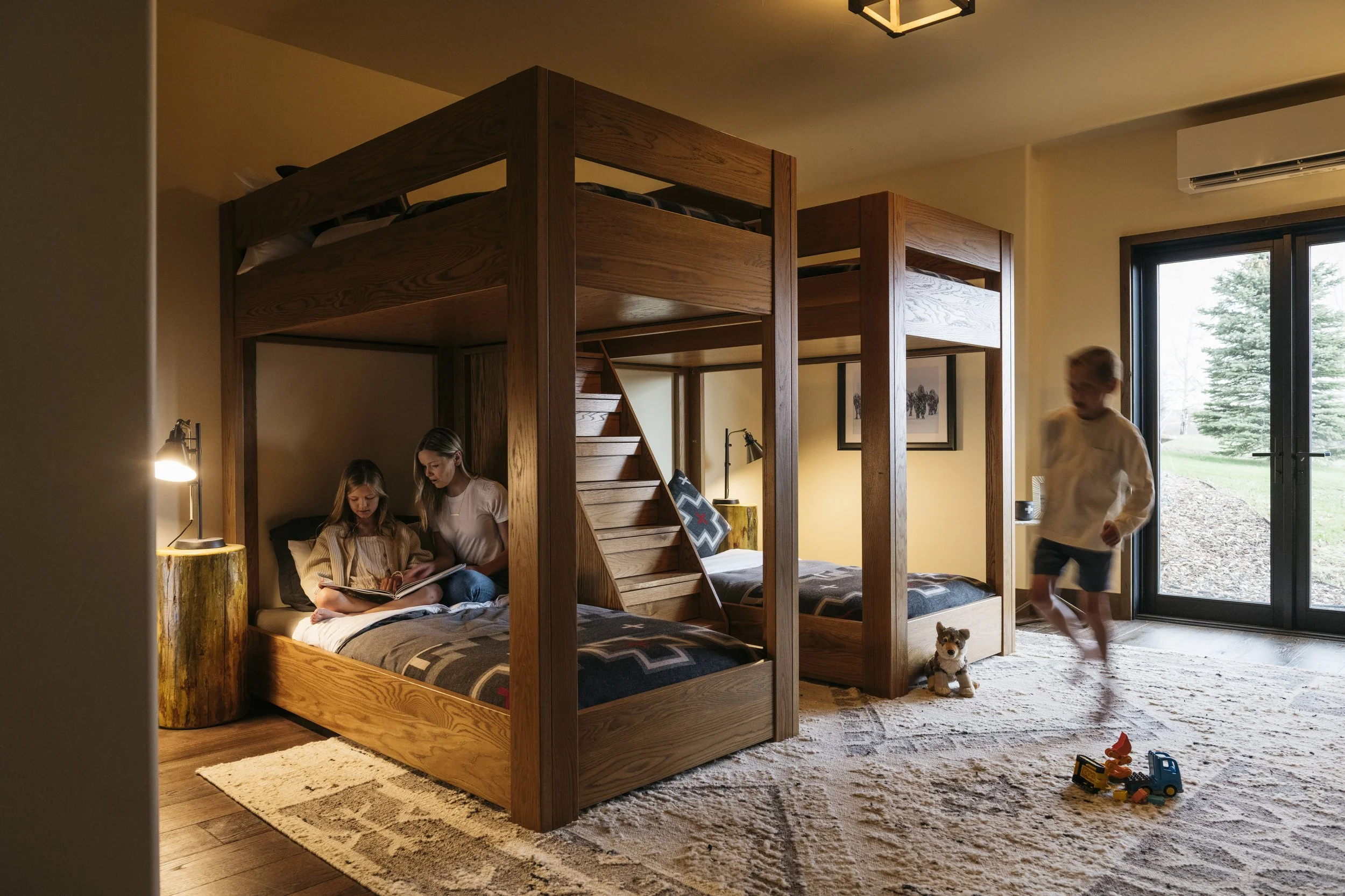 A family of children playing and reading in a bedroom with a wooden bunk bed, large glass doors showing greenery outside, and a plush carpet.