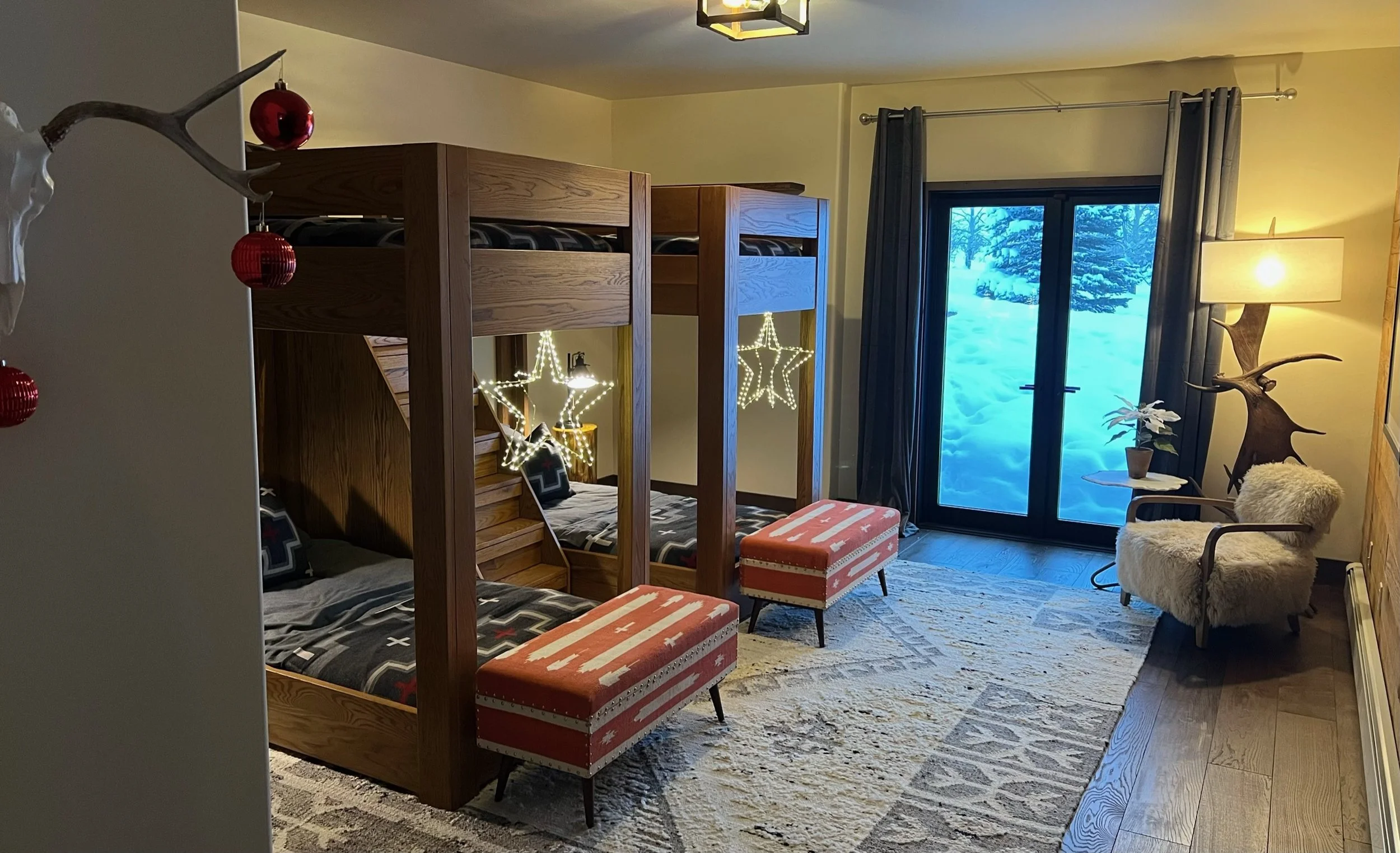 Interior of a cozy mountain room with a wooden bunk bed, two patterned stools, a fireplace with star-shaped lights, a plush armchair with a sheepskin cover, a large window showing a snowy landscape outside, and holiday decorations including a mounted