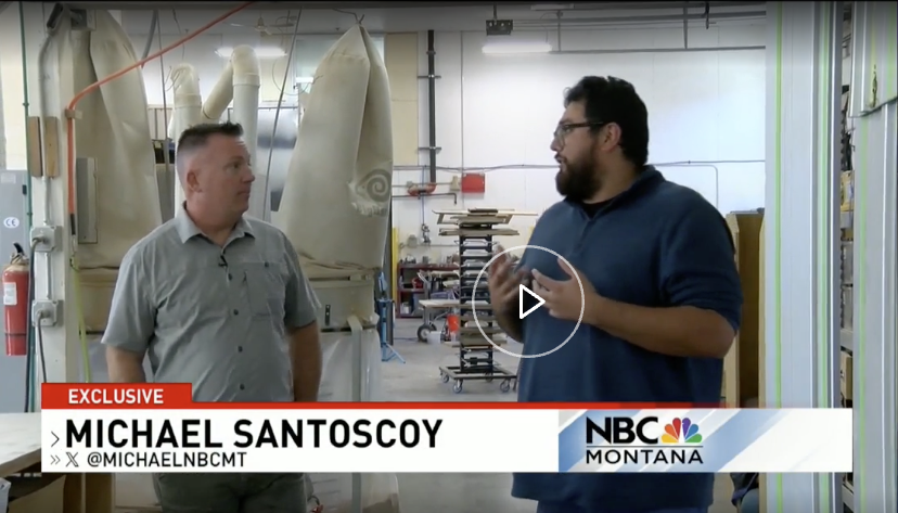Two men having a conversation in an industrial workspace with large fabric or paper rolls hanging from the ceiling and various equipment in the background.