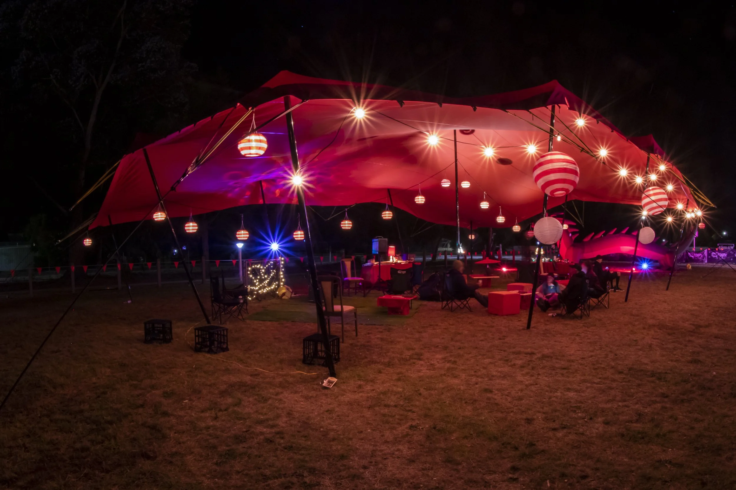 Outdoor nighttime event with seating under a large red canopy, decorated with red and white paper lanterns and string lights, featuring people sitting at tables and chairs.