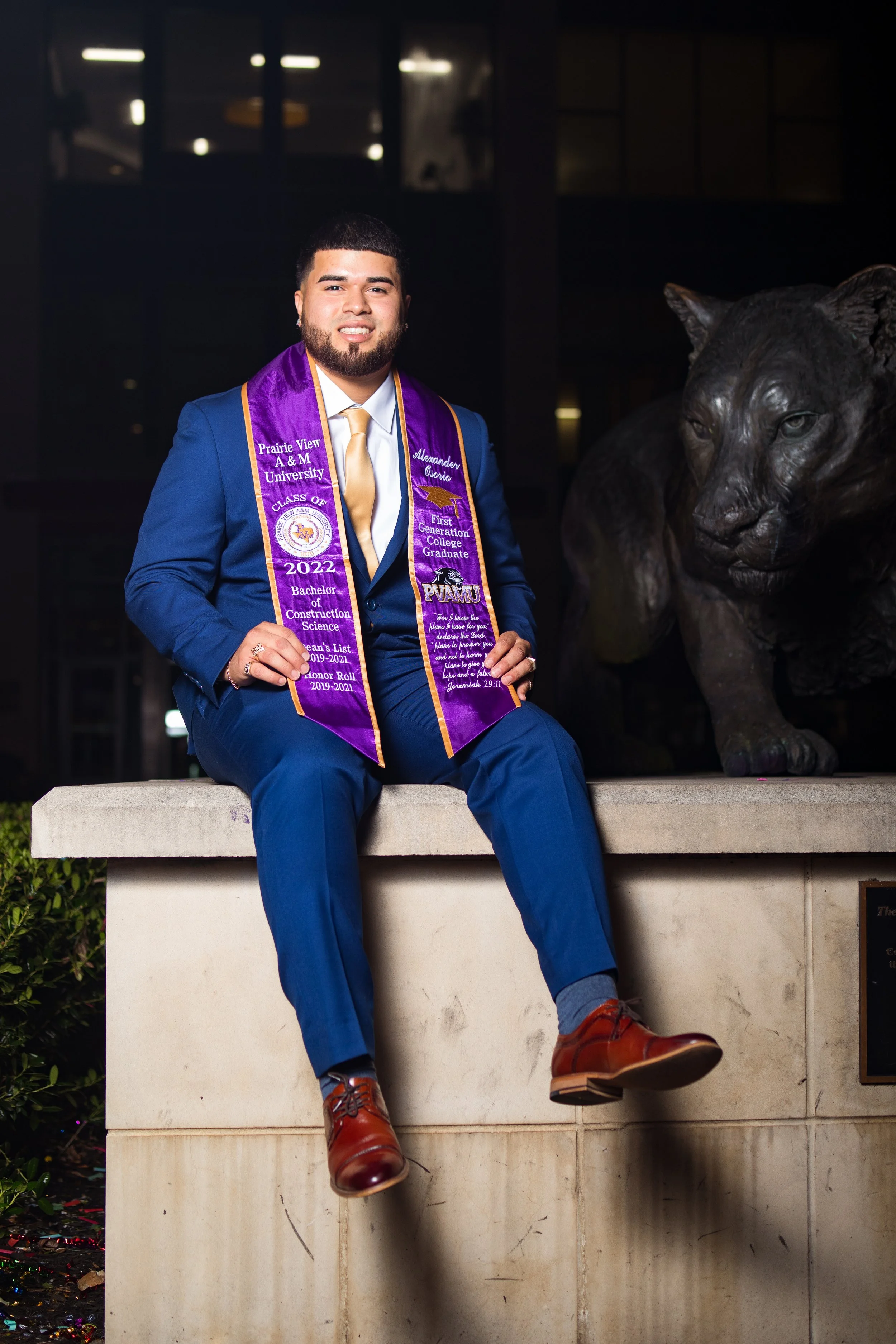 Young man in a blue suit with a gold tie sitting on a concrete ledge next to a lion statue, wearing a purple graduation stole from Prairie View A&M University.