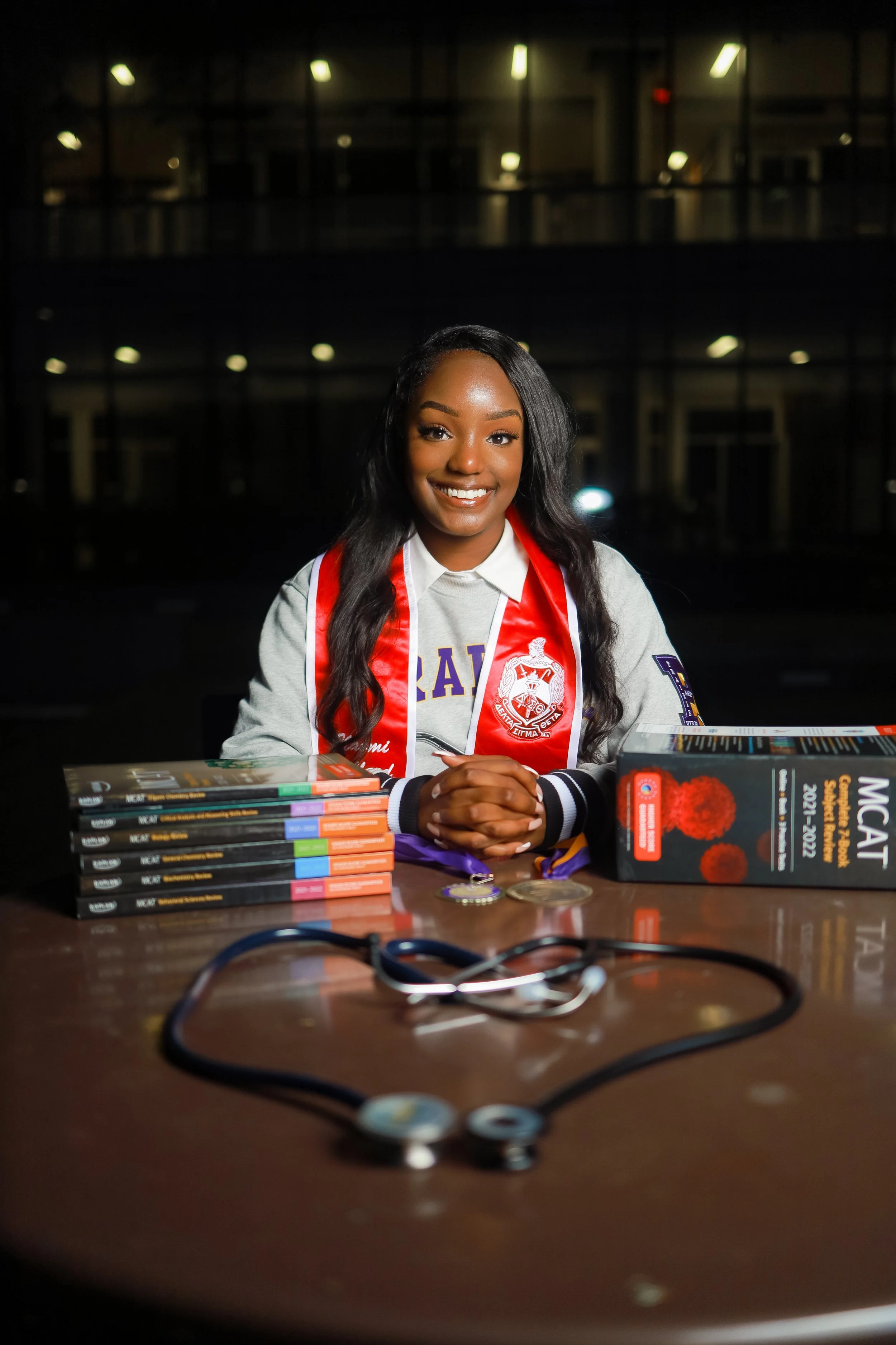 A young woman wearing a graduation gown and cap, sitting at a table with medical textbooks, a stethoscope, and medals, in an indoor setting at night, smiling at the camera.