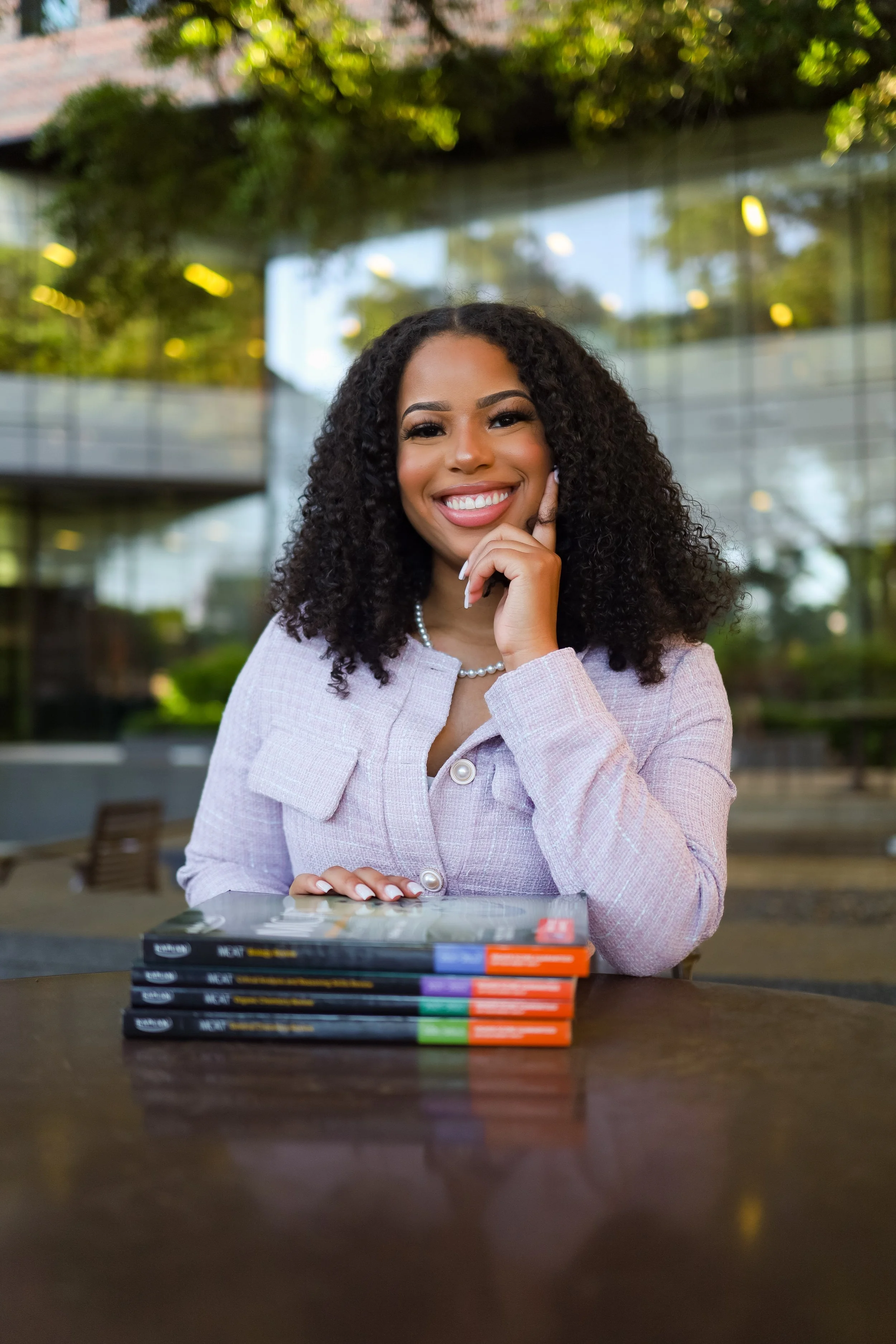 A smiling woman with curly hair, wearing a light purple blazer, sitting at a table with stacked magazines, outdoors with a modern glass building and trees in the background.