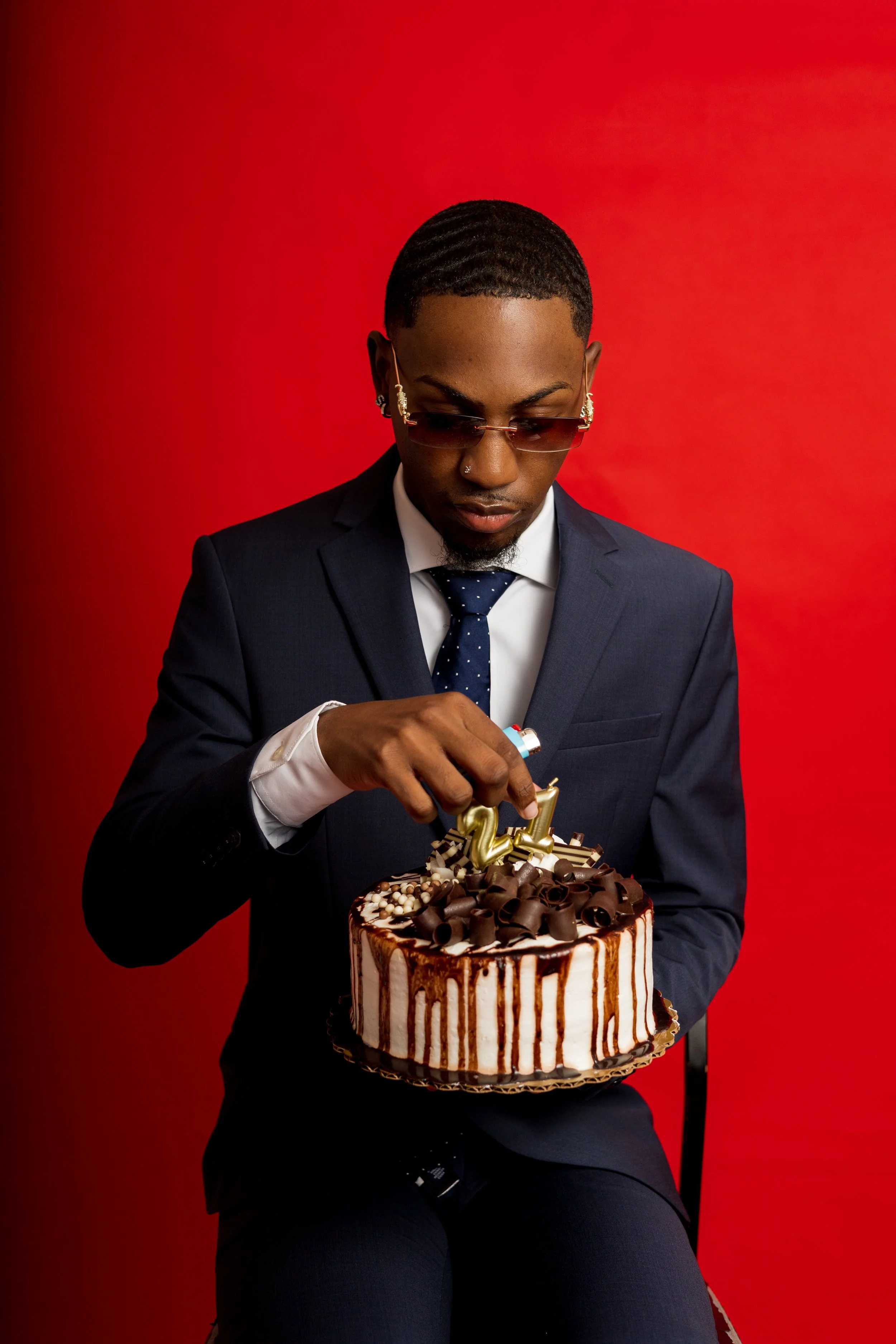 A man in a blue suit, white shirt, and polka dot tie is sitting against a red background, holding a birthday cake and wearing sunglasses.