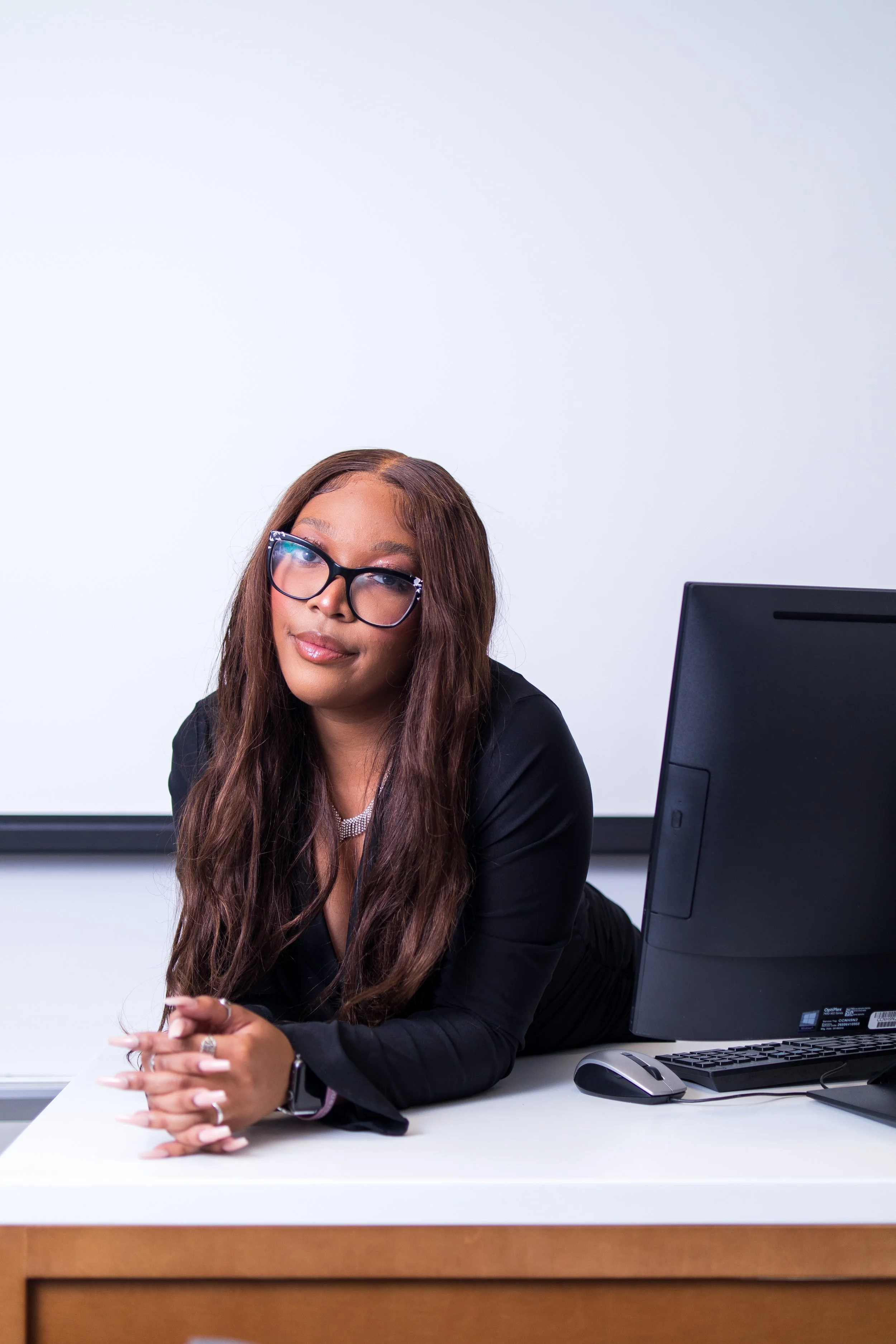 A woman with long brown hair wearing glasses, a black blazer, and a silver necklace, sitting at a desk with her hands clasped, in front of a computer monitor, in a professional setting.