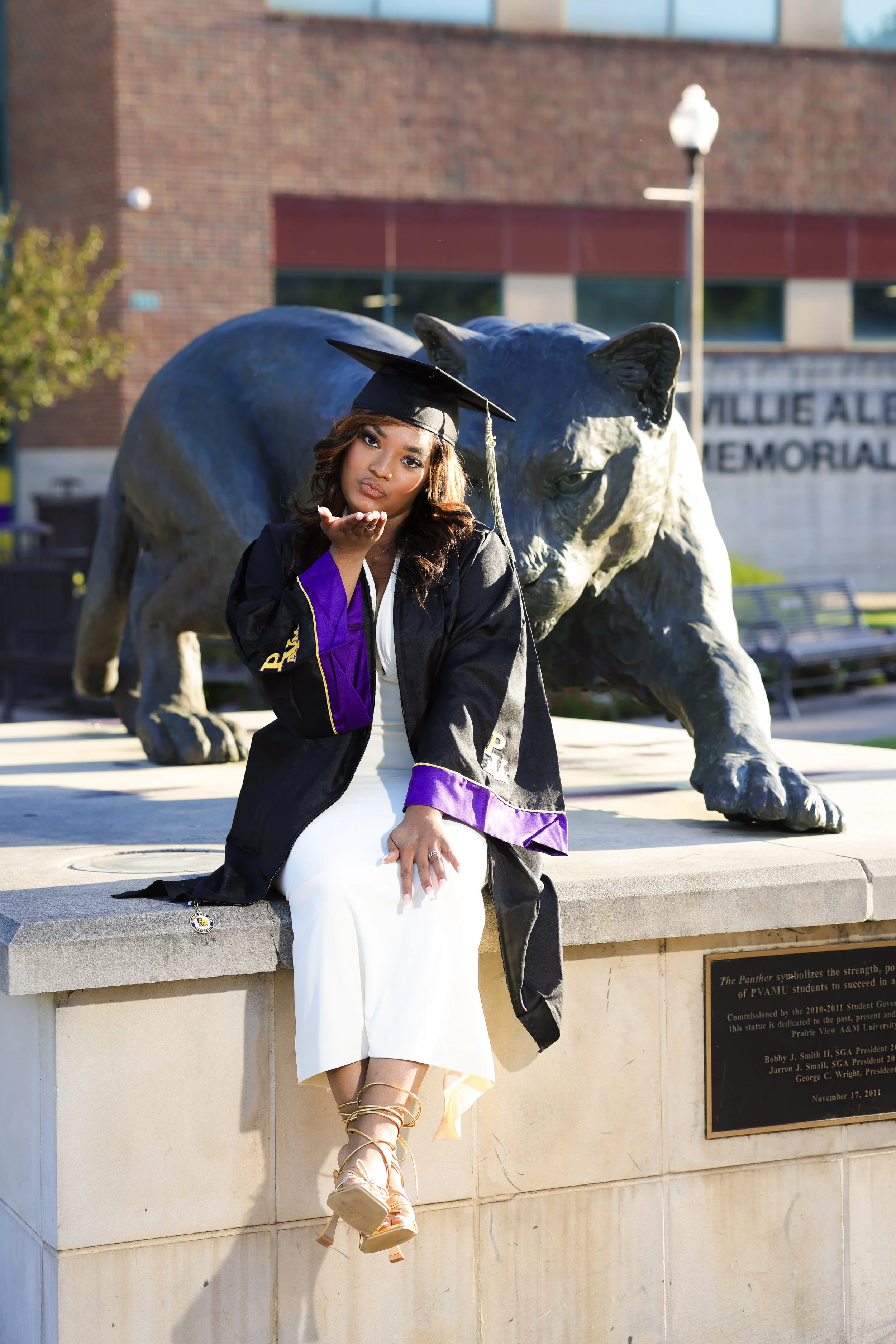 A young woman in a graduation cap and gown sits on a monument featuring a panther, blowing a kiss towards the camera. She wears a black gown with purple accents and beige high-heeled lace-up sandals. The background shows a brick building and a statue