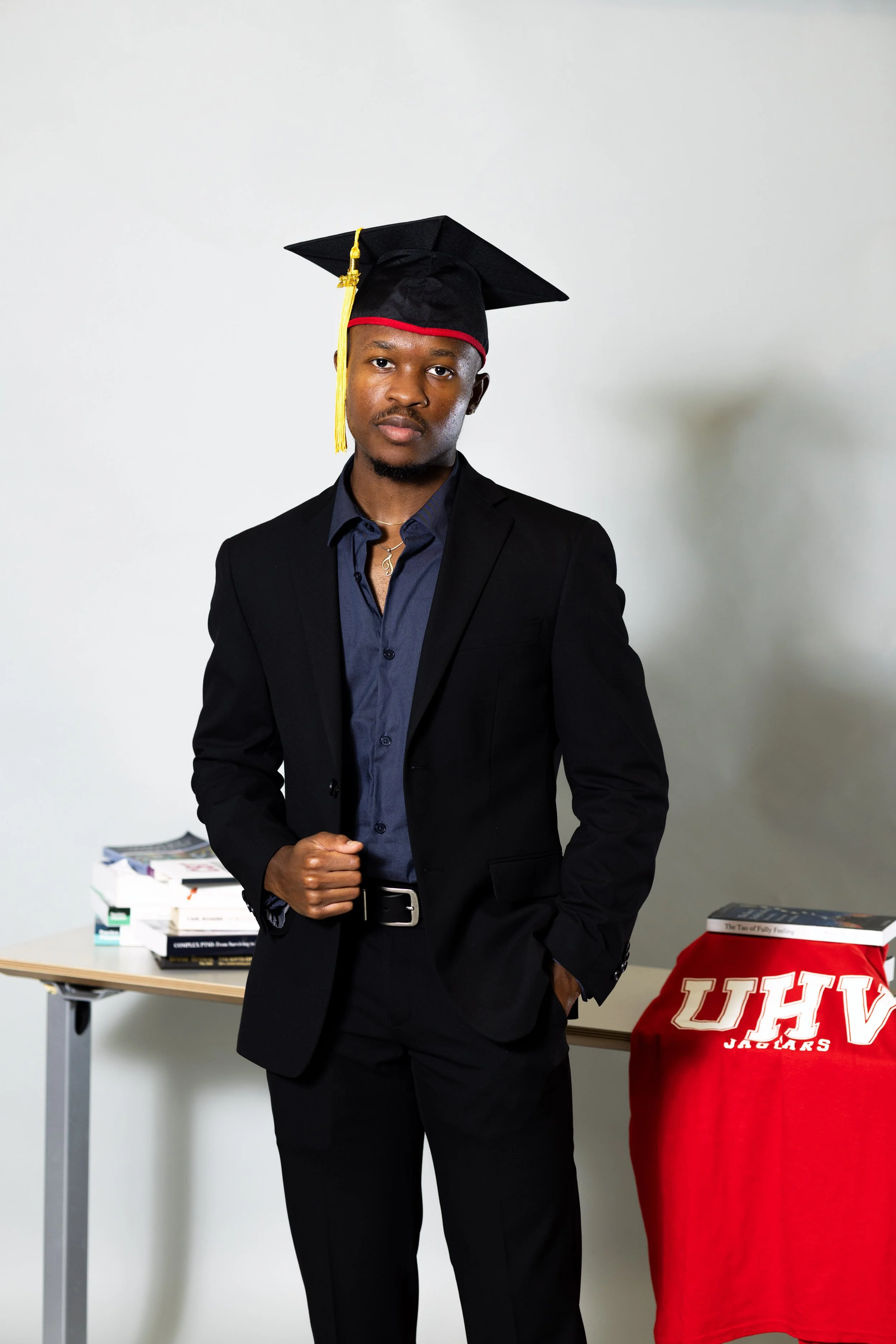 A young man in a black suit and blue shirt wearing a graduation cap stands in front of a table with books and a red cloth with white letters UHV. He has a serious expression and one hand is clenched into a fist while the other is in his pocket.
