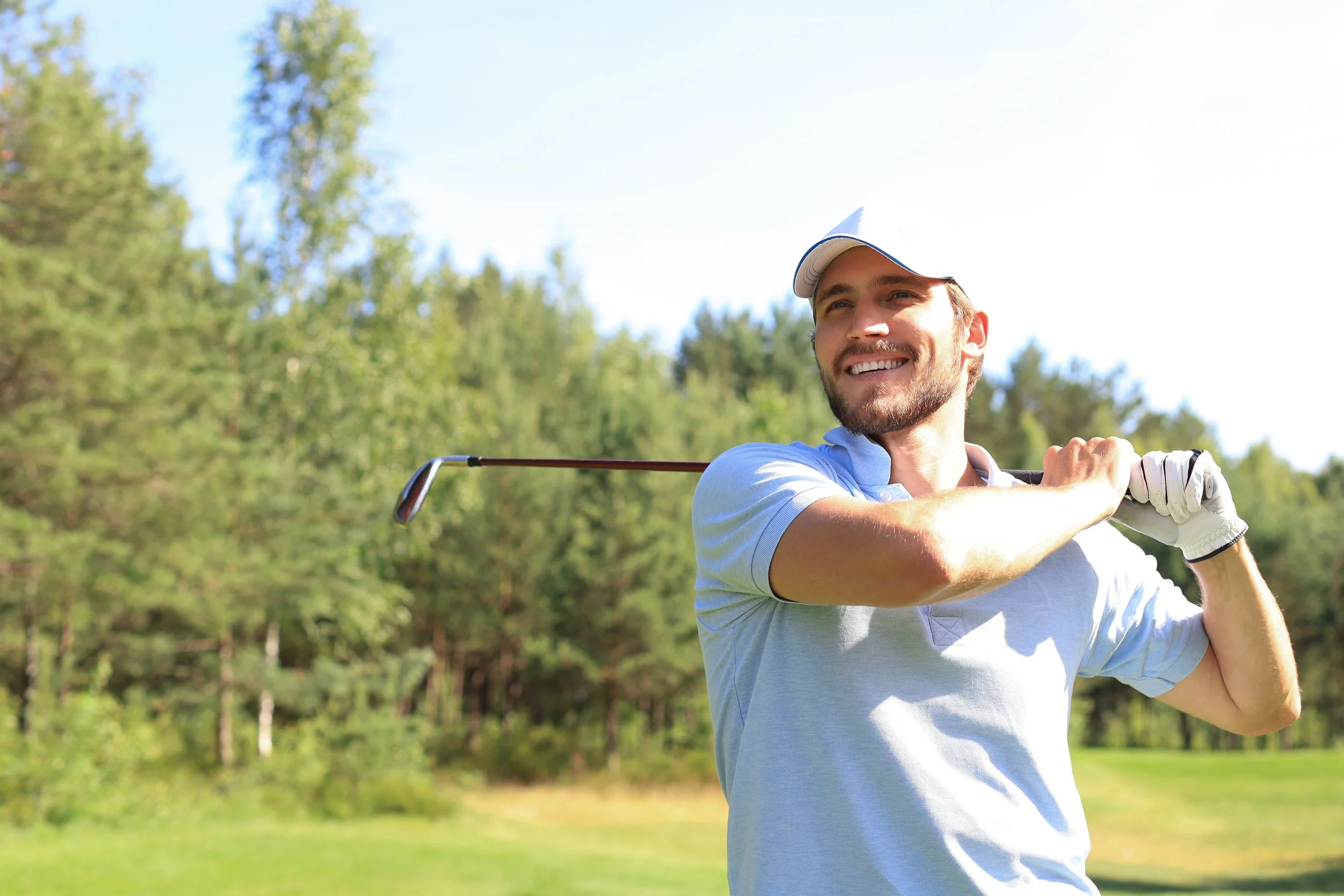 A smiling man in golf attire holding a golf club on a golf course with trees in the background.