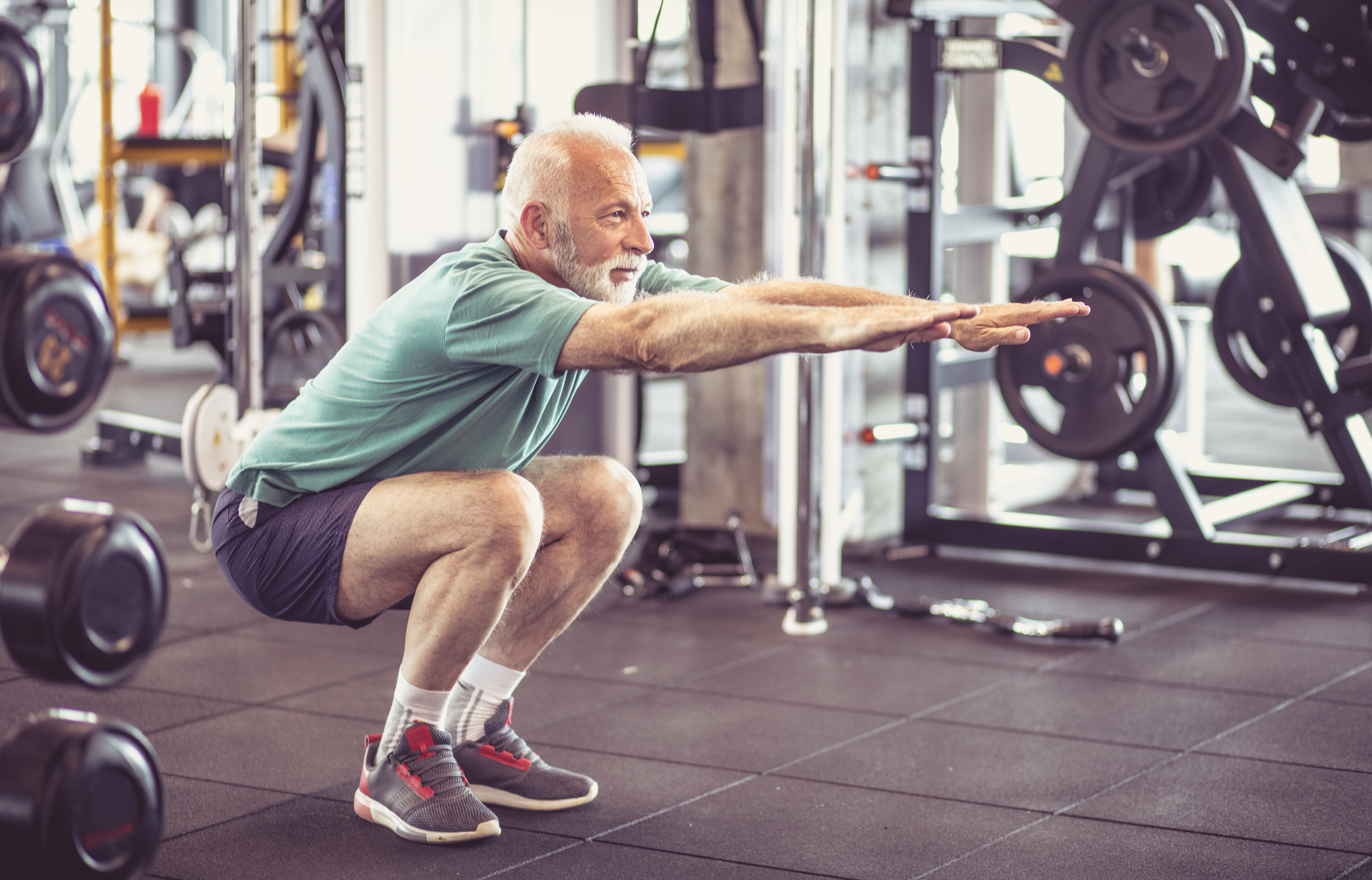 An elderly man with grey hair and beard performing a squat exercise in a gym, wearing a green shirt, black shorts, and grey sneakers with red accents.