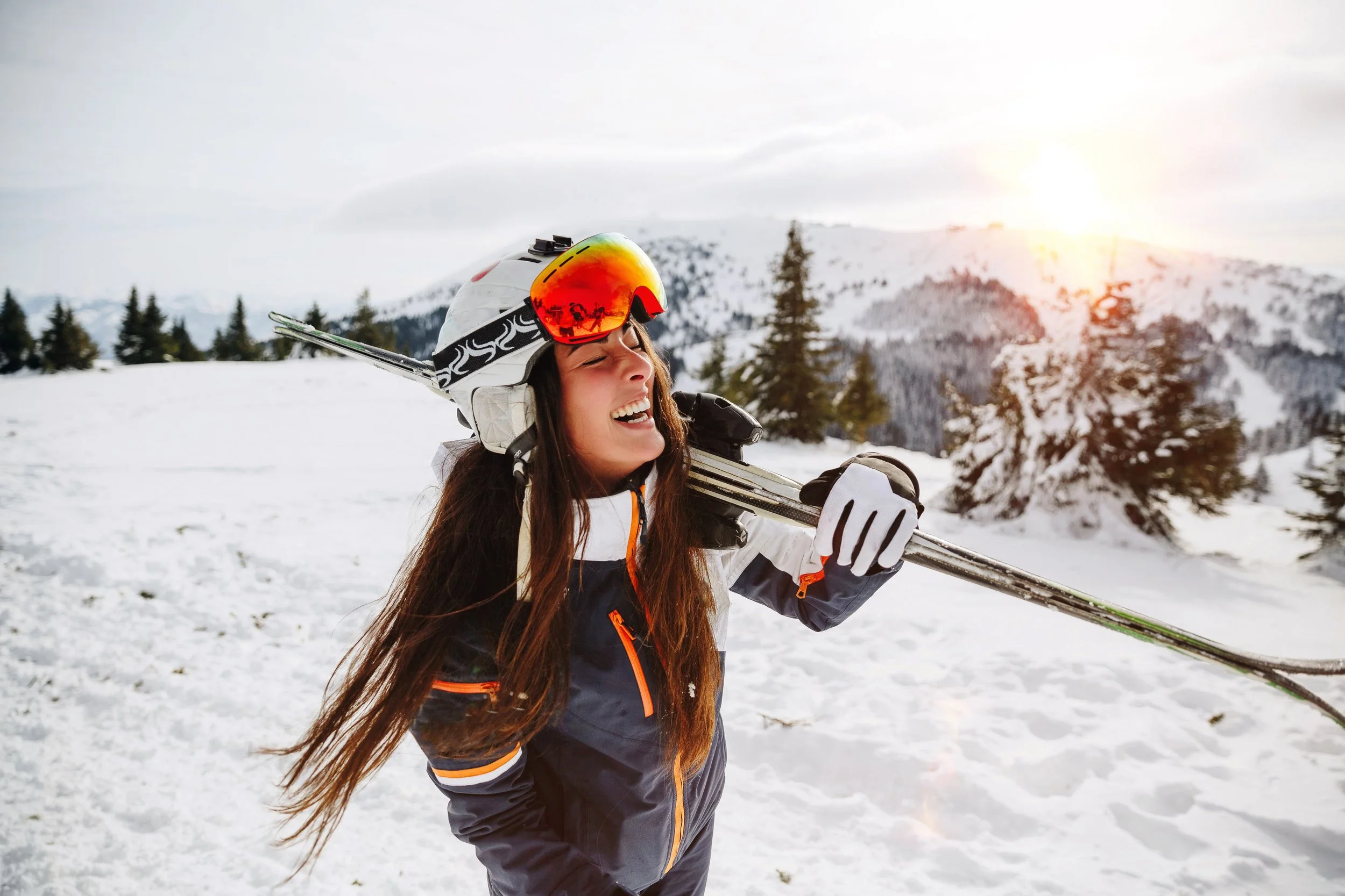 A woman in snow gear carrying skis on her shoulder, smiling with her eyes closed, in a snowy mountain landscape at sunset.