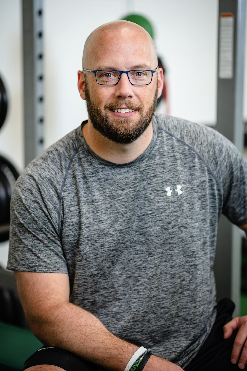A man with a beard, glasses, and a shaved head wearing a gray Under Armour T-shirt, sitting in a gym.