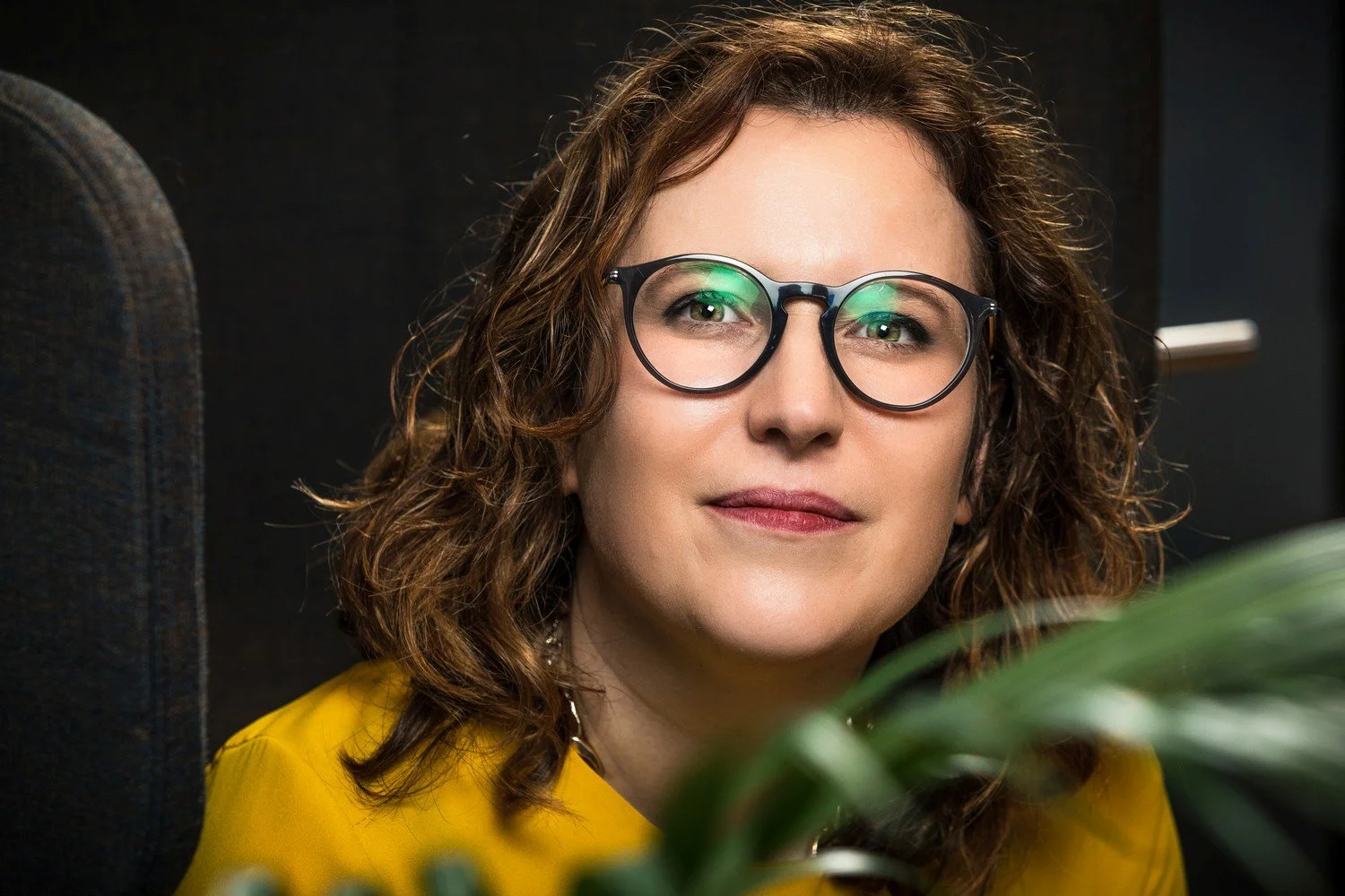 Close-up of a woman with curly brown hair wearing round glasses and a yellow top, sitting in a dark indoor setting with a blurred plant in the foreground.