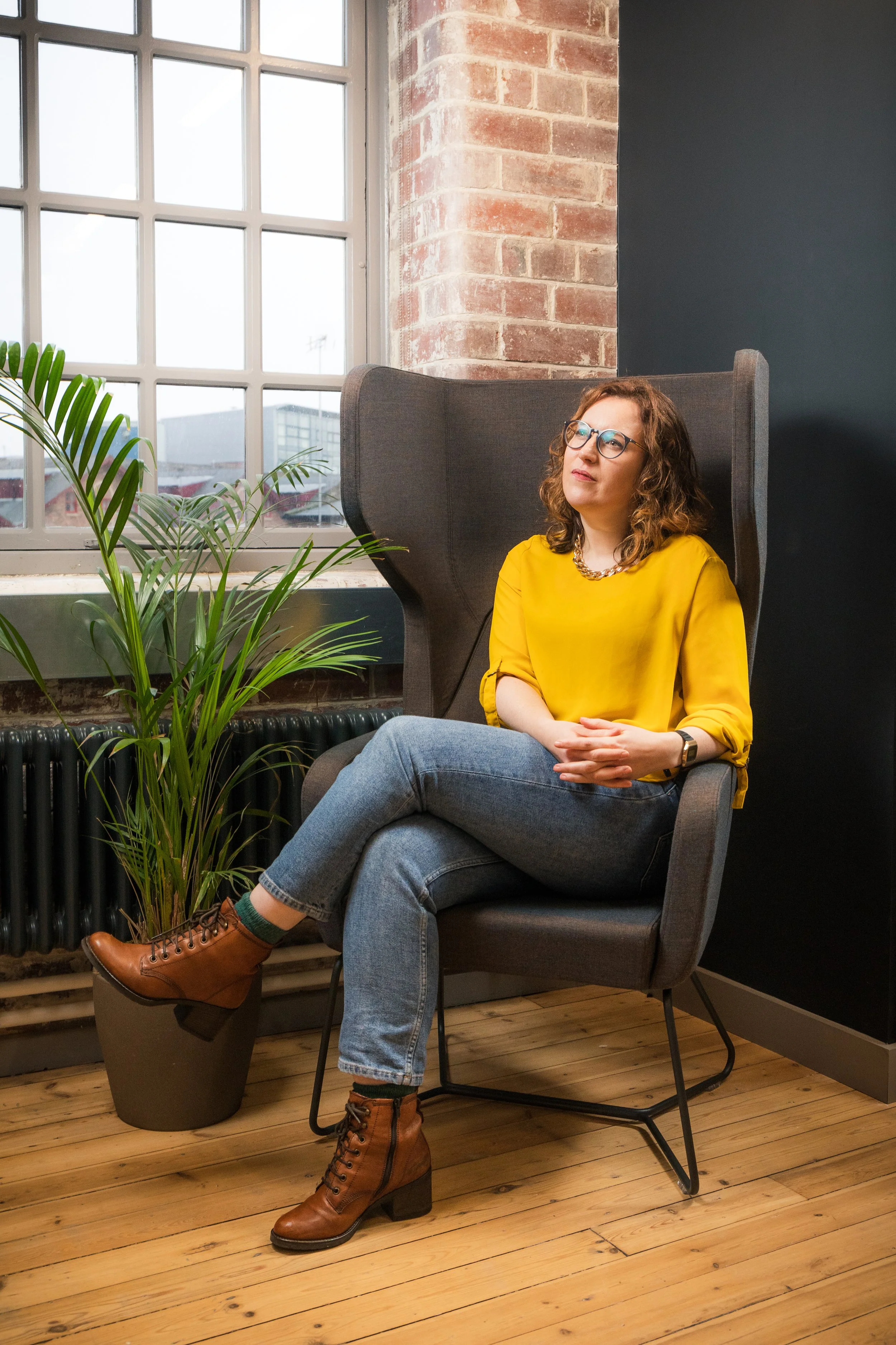 A woman with curly brown hair, glasses, and a yellow blouse is sitting in a modern, high-back gray chair next to a window in an industrial-style room with exposed brick walls and a wooden floor. She is sitting with her legs crossed and looking thoughtfully ahead.