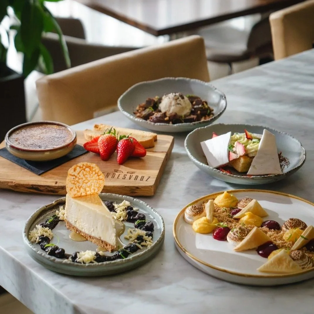 Assorted desserts on a table including a cheesecake with blueberries, a fruit tart, a variety of plated sweet treats, fresh strawberries, and baked goods, in a cozy restaurant setting.